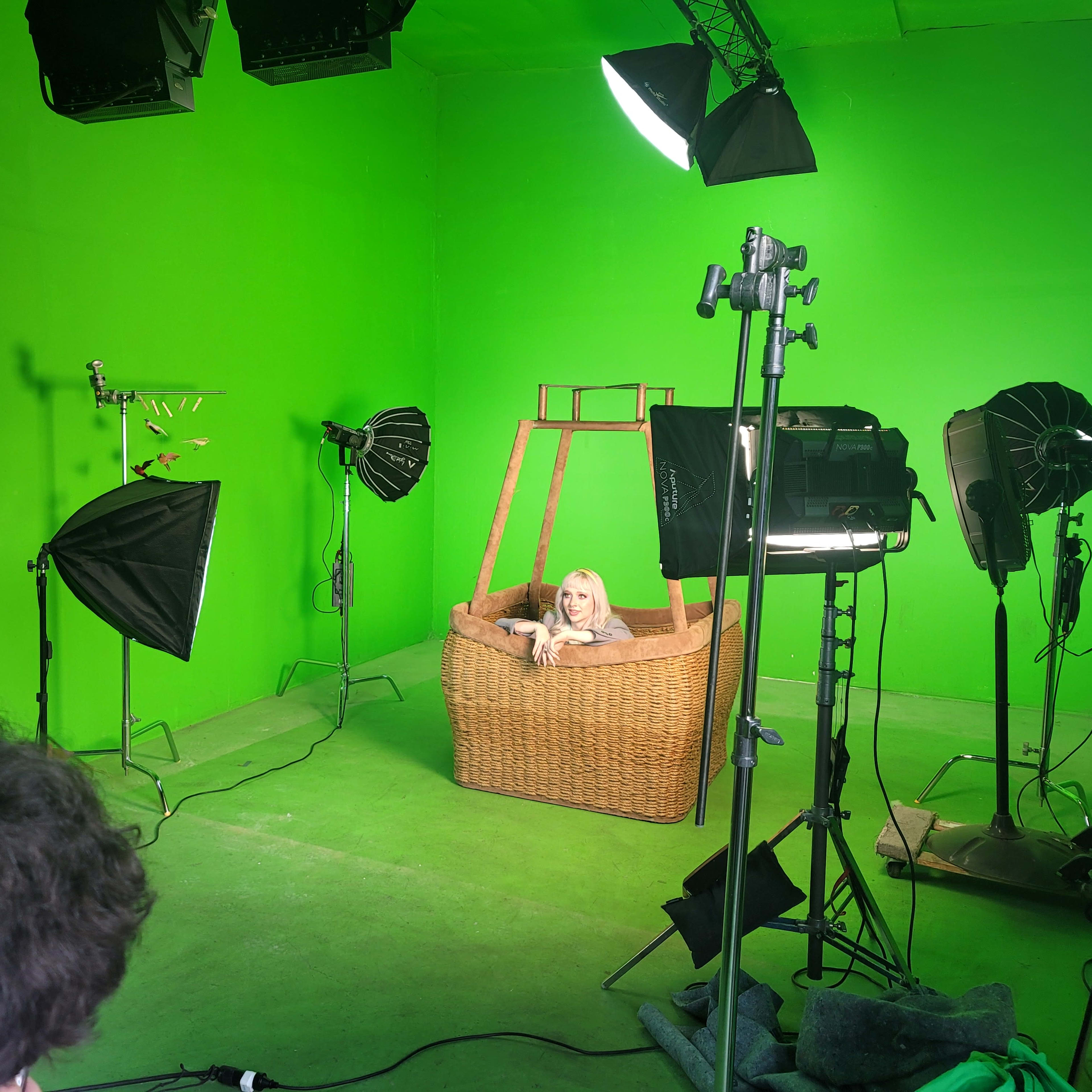 The scene shows a model sitting in a wicker basket surrounded by studio lighting in a green-screen room.