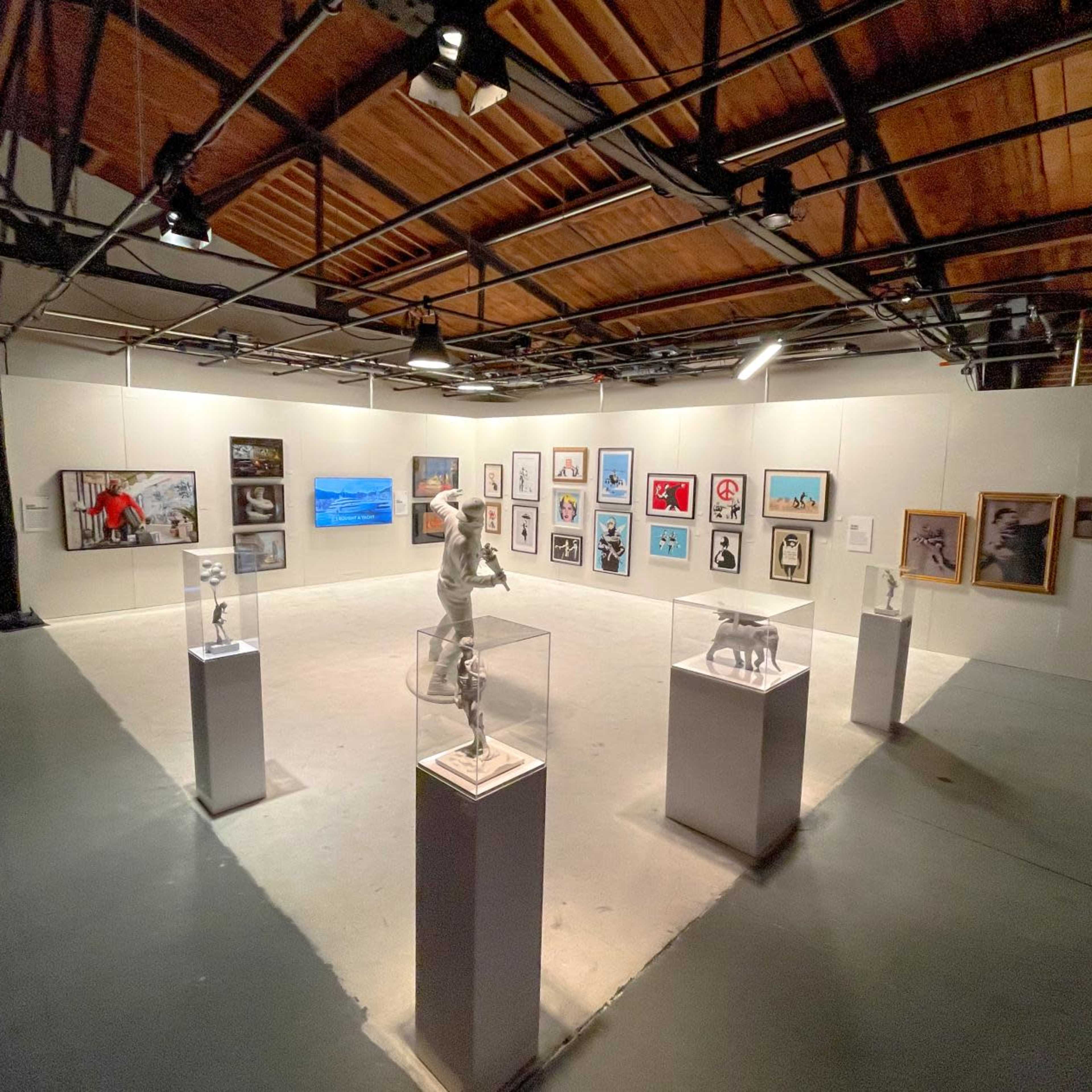 A contemporary art gallery featuring various framed artworks and sculptures arranged in a spacious, well-lit room with high ceilings.