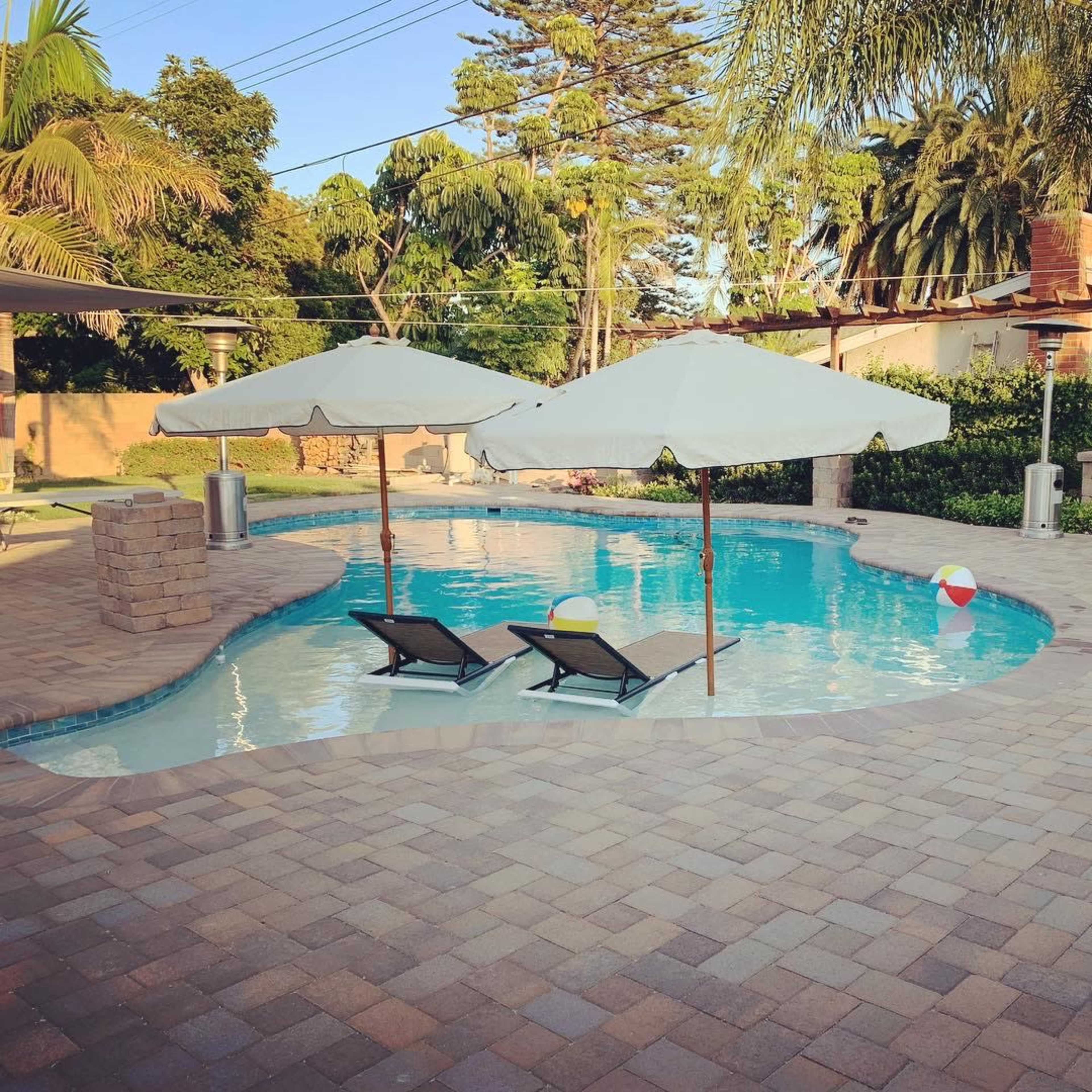 A pool area featuring two lounge chairs under umbrellas, surrounded by palm trees and a paved patio with a beach ball nearby.