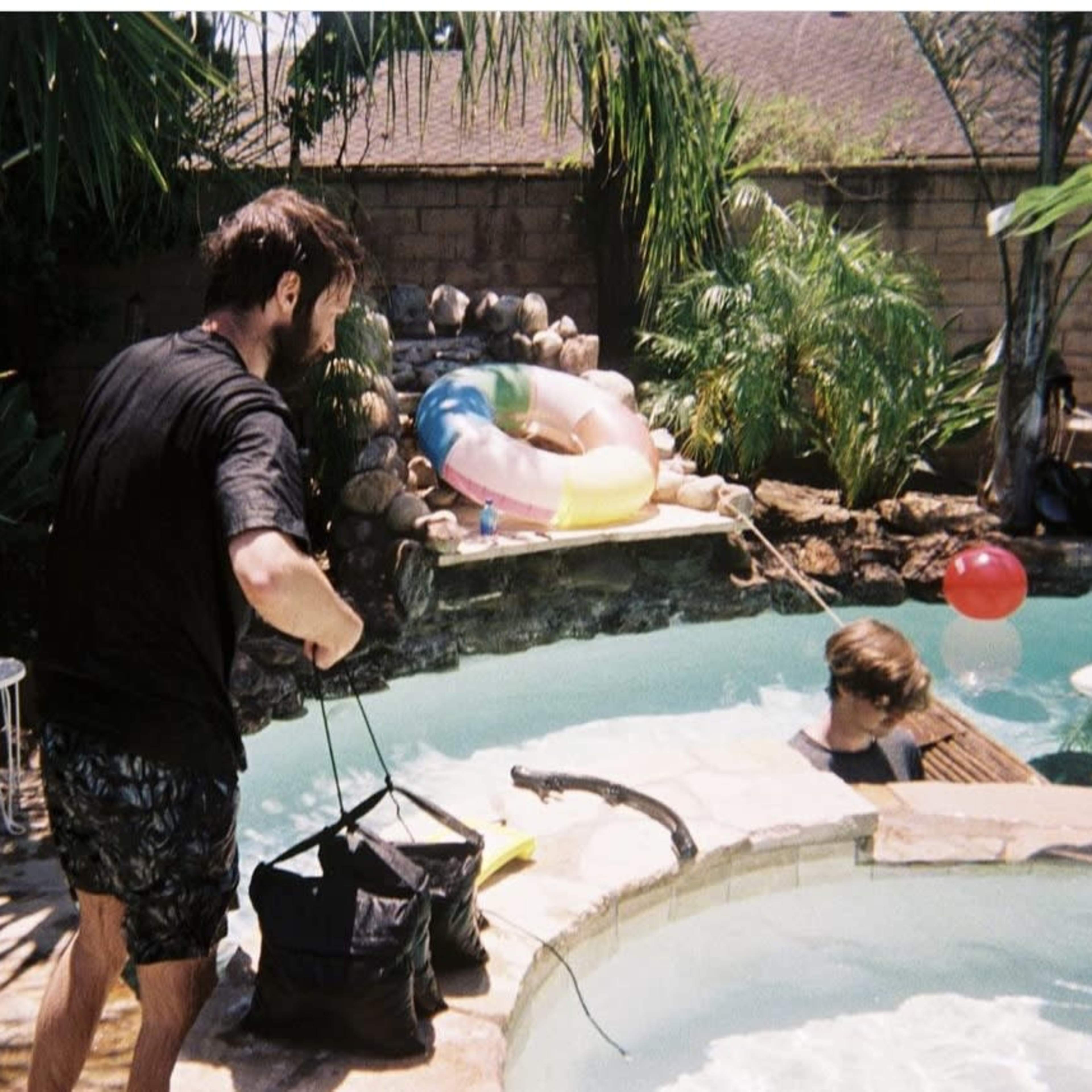 Two people are near a backyard pool surrounded by tropical plants, with one person on the pool's edge and the other in the water.