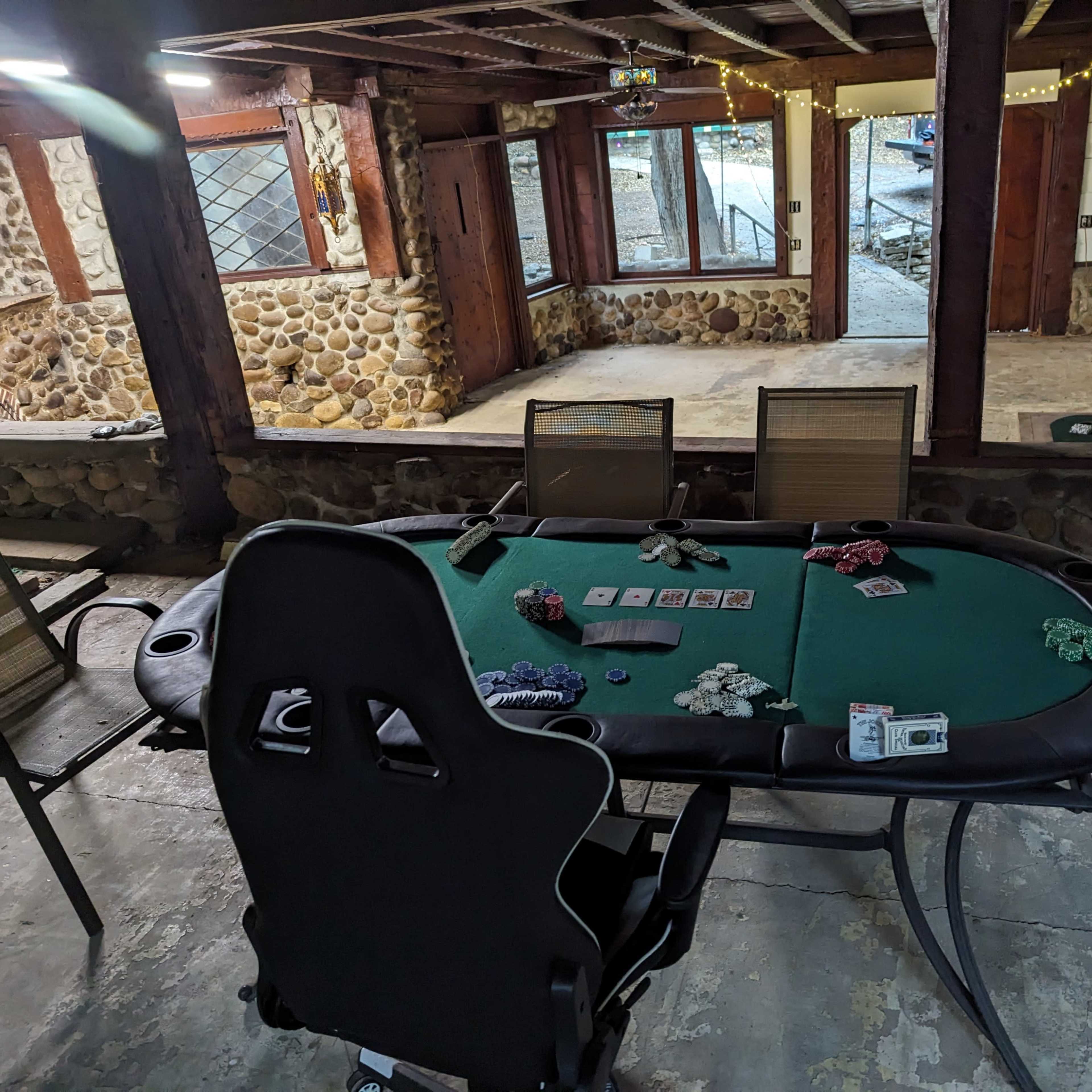 A poker table with poker chips and cards is set up in a rustic room with stone walls and large windows.