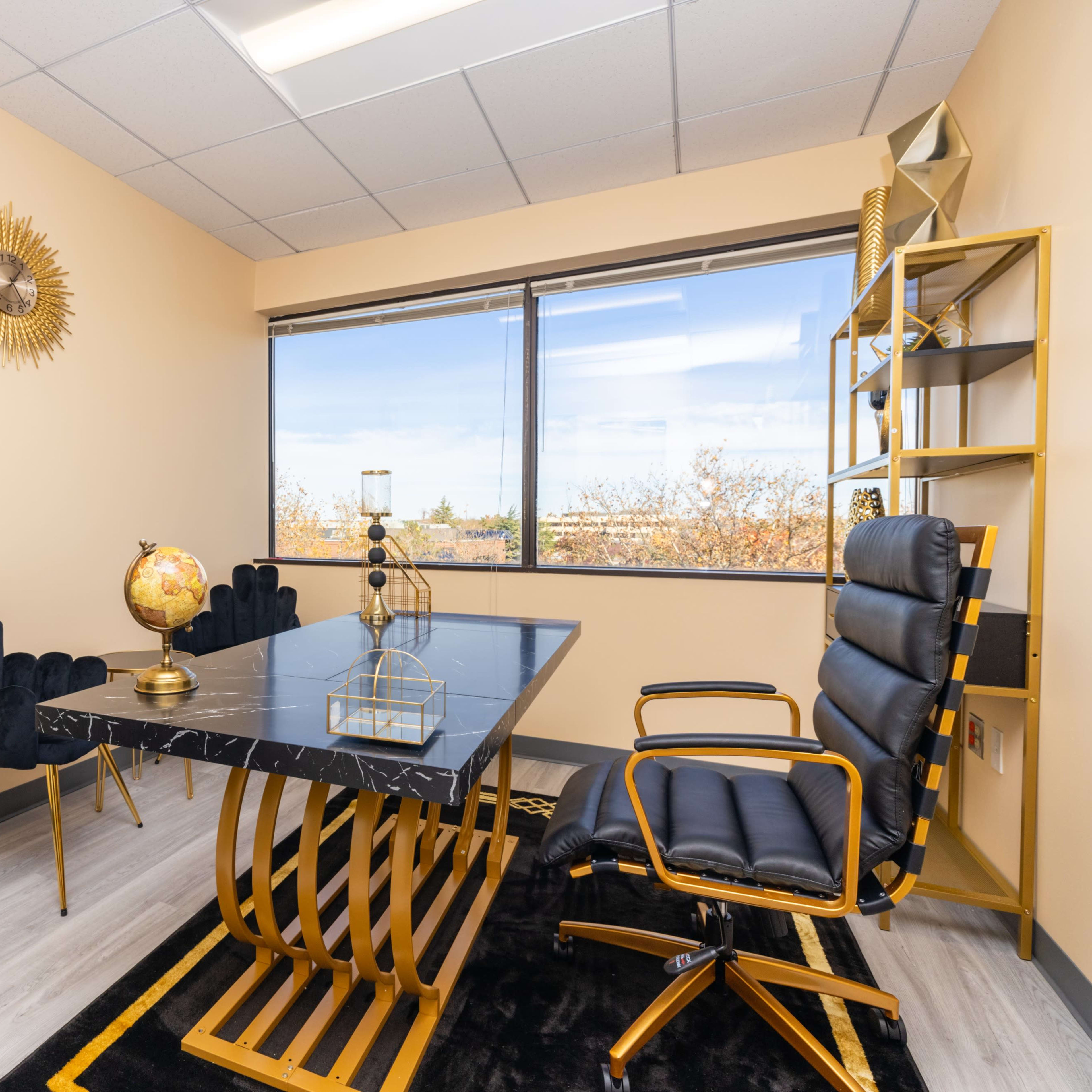 A modern office space featuring a black marble table, a black leather chair, two upholstered chairs, and a decorative globe, all set against a light-colored wall and large window.