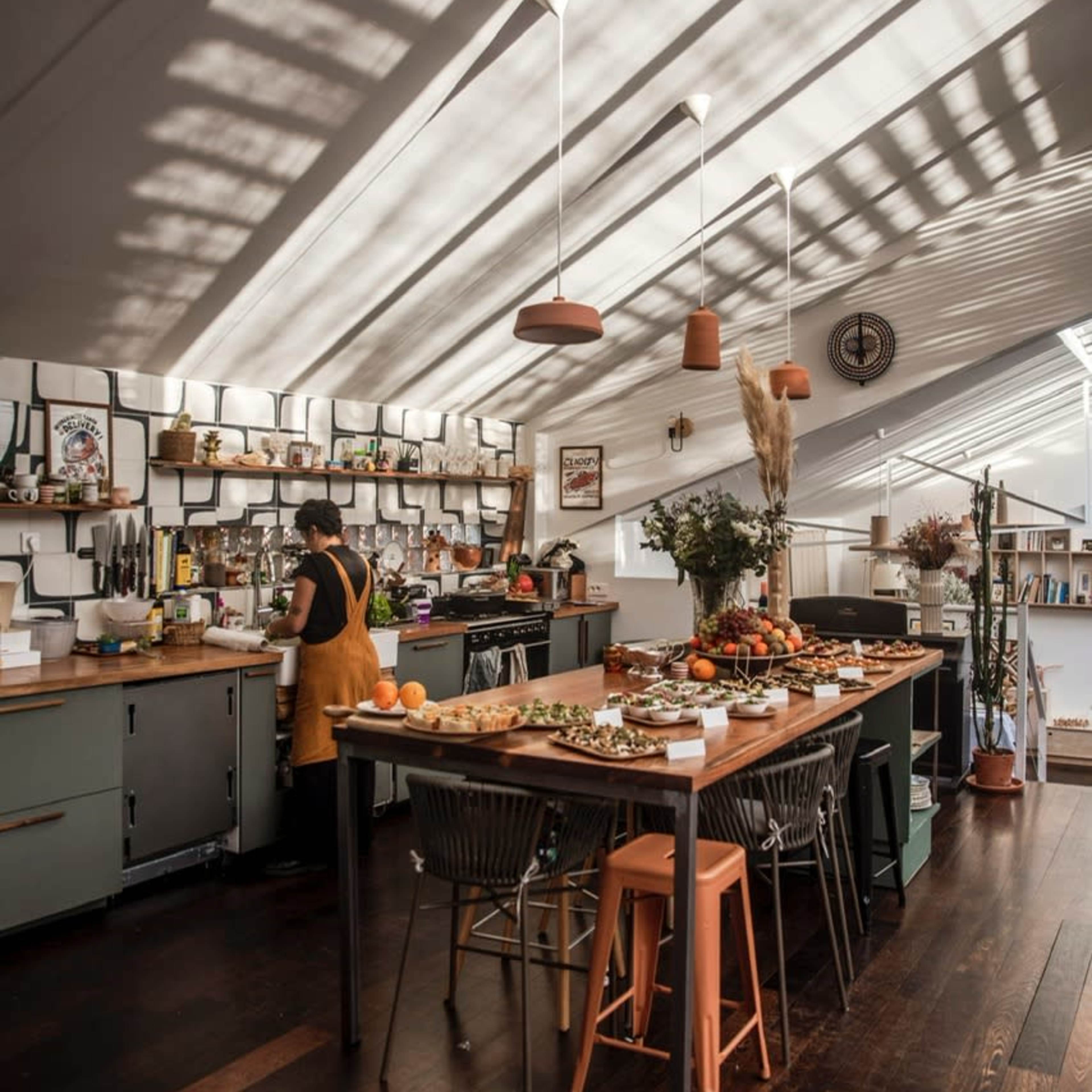 A spacious kitchen features a large wooden island covered with various dishes, while sunlight streams through skylights, casting shadows on the walls and floor.