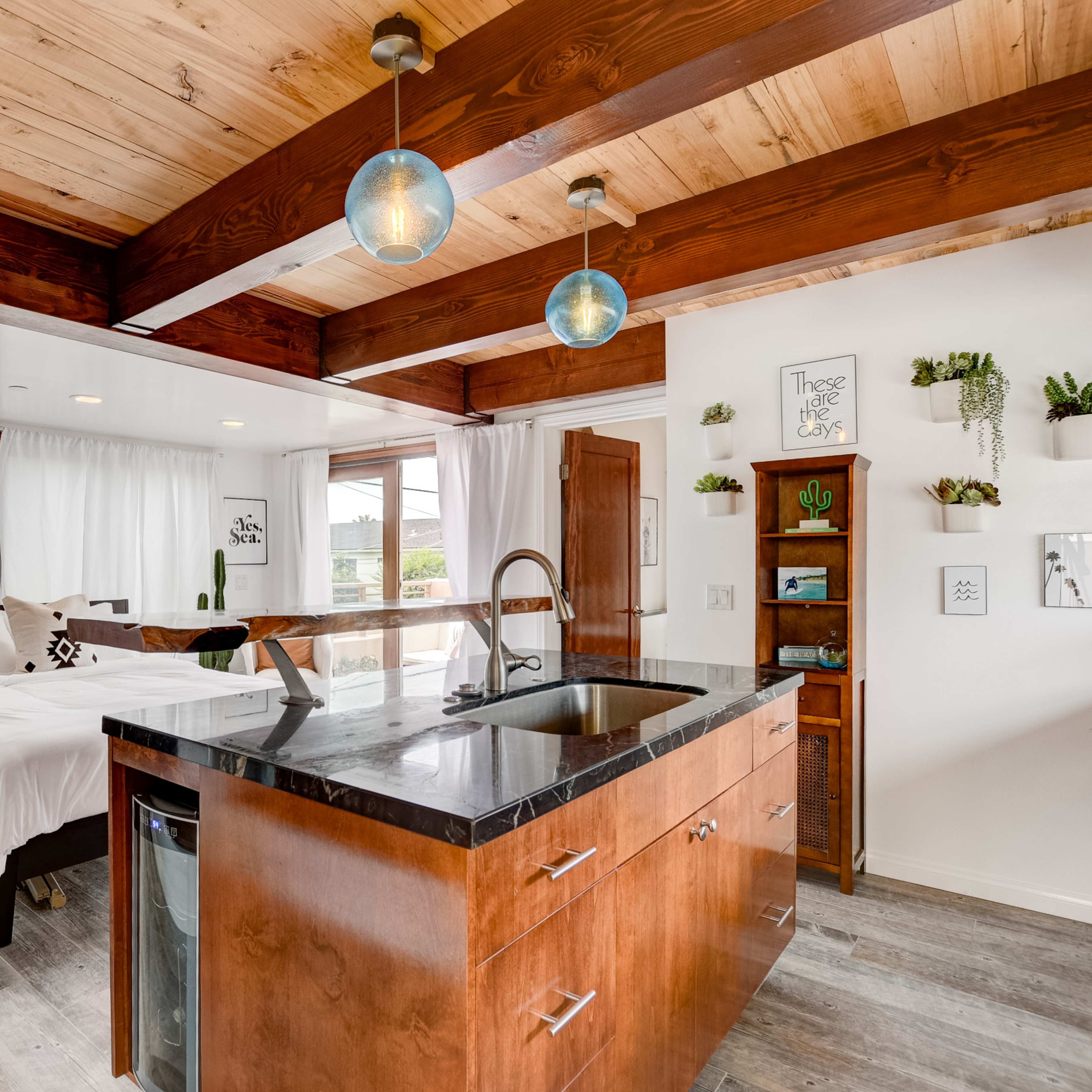 A modern studio apartment with wooden beams, a kitchen island, and a bed area separated by sheer curtains.