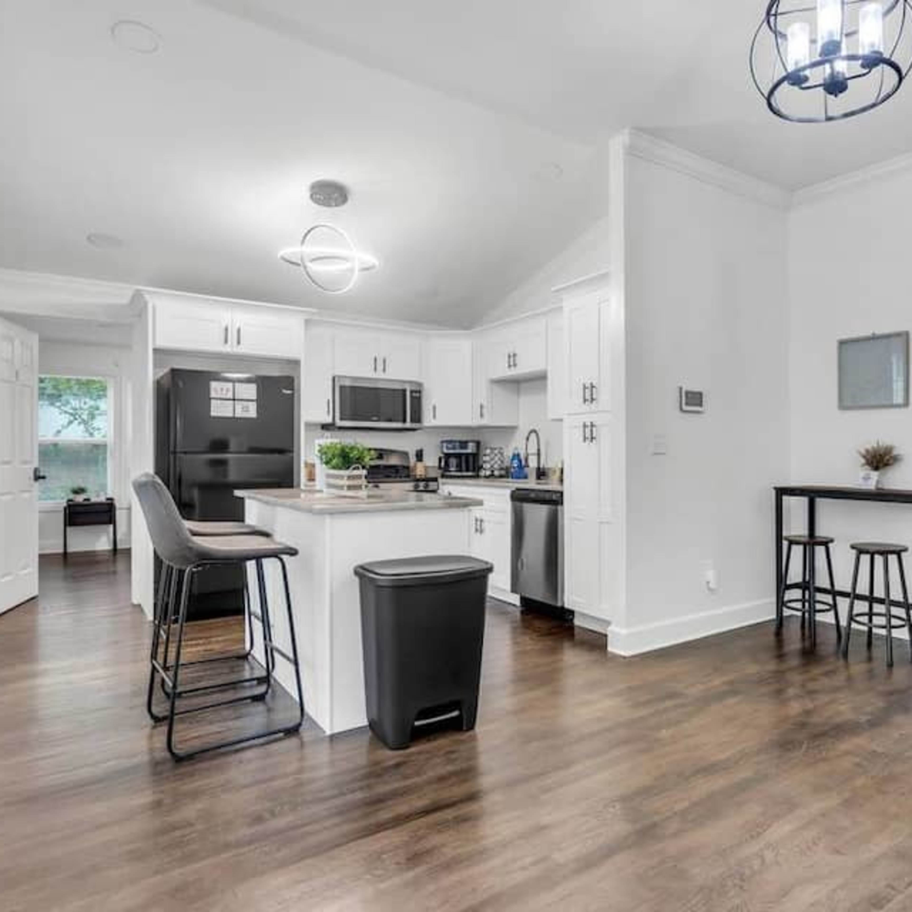 A modern kitchen area with white cabinetry, stainless steel appliances, a breakfast bar with stools, and a small dining table in an open layout.