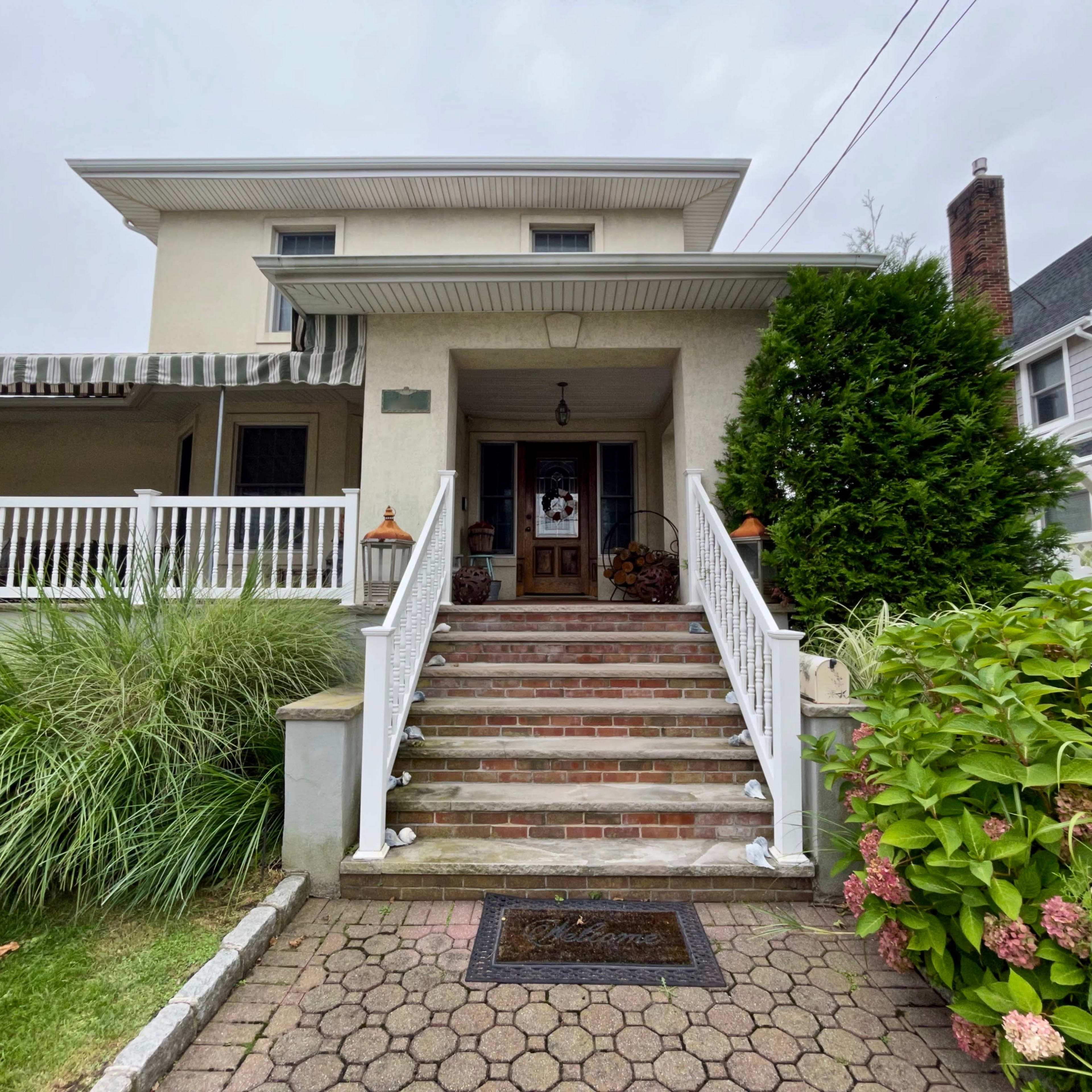A two-story house with a front porch and steps leading to the entrance, surrounded by shrubs and flowers.