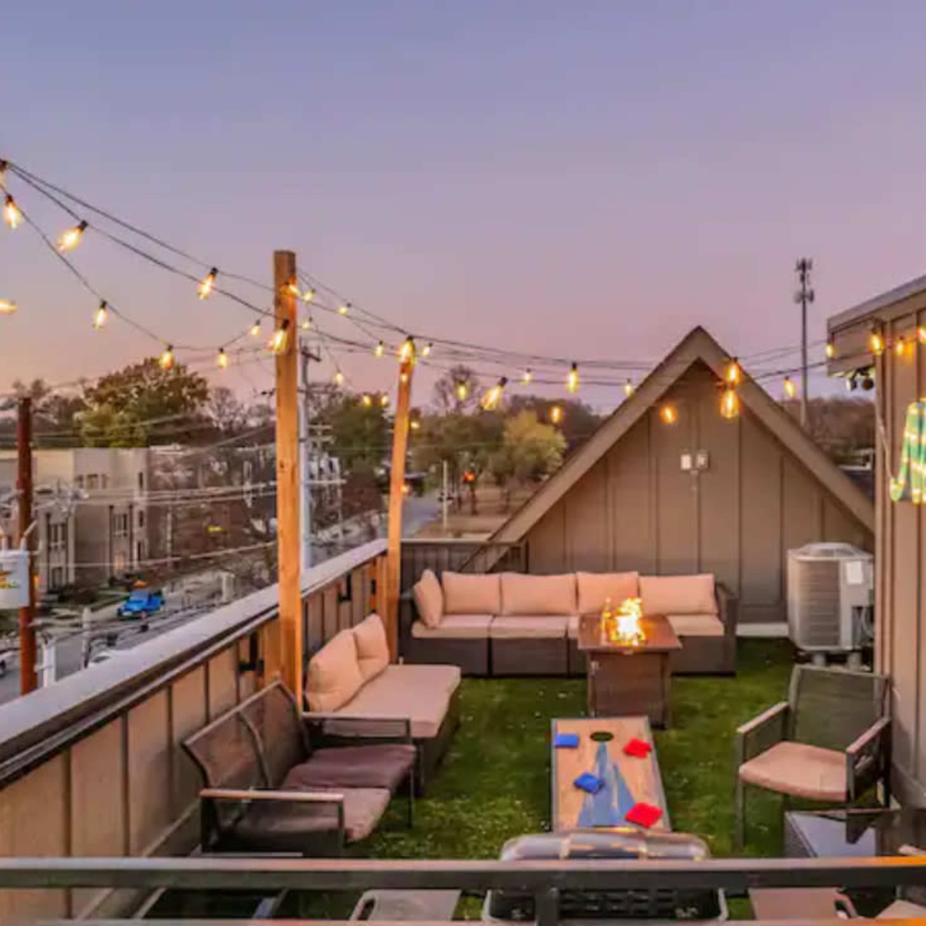 A rooftop terrace with seating arrangements, string lights, and a small fire pit, set against a twilight sky.