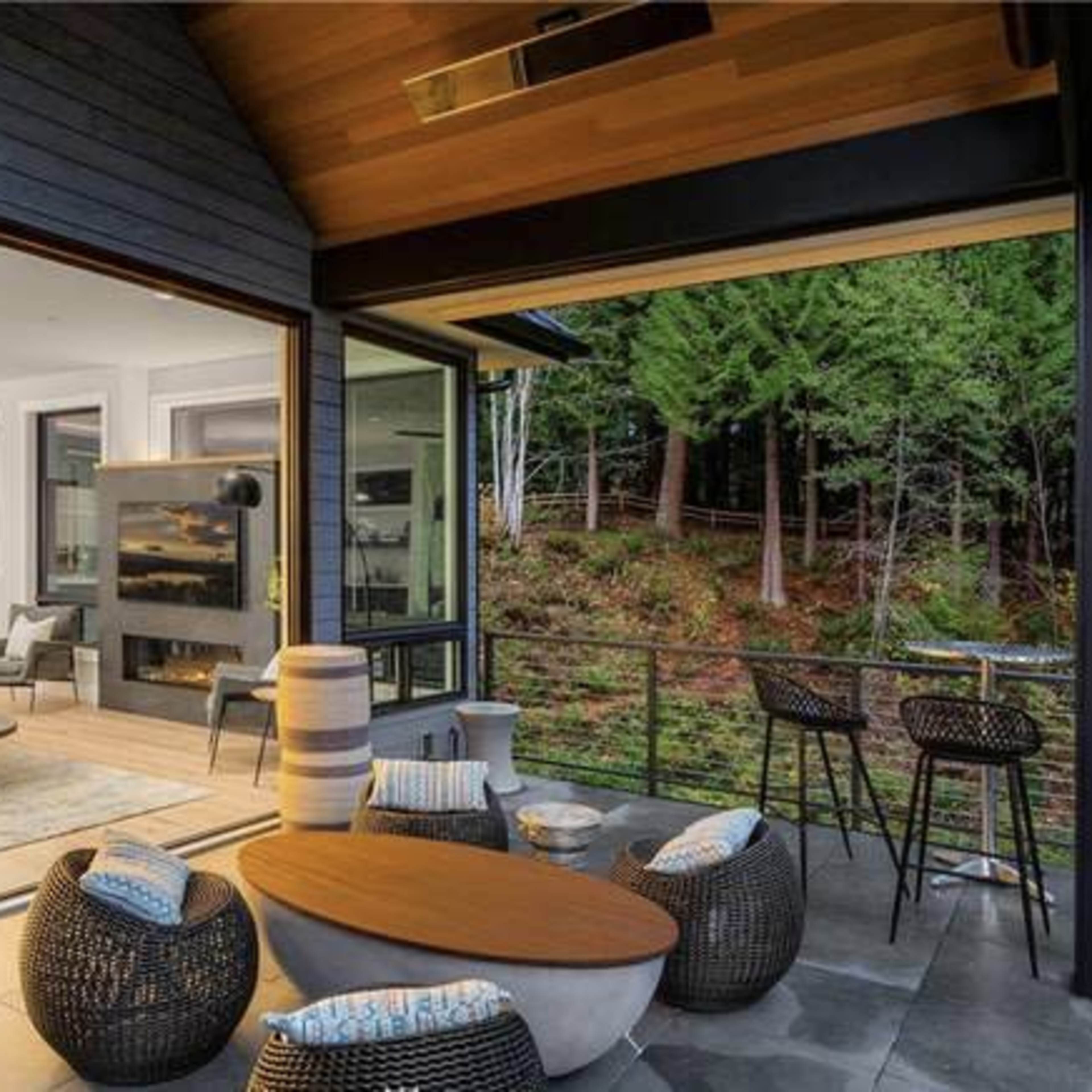 A modern outdoor seating area features a round table surrounded by wicker chairs, overlooking a lush forest through large glass doors that connect to an interior living space.