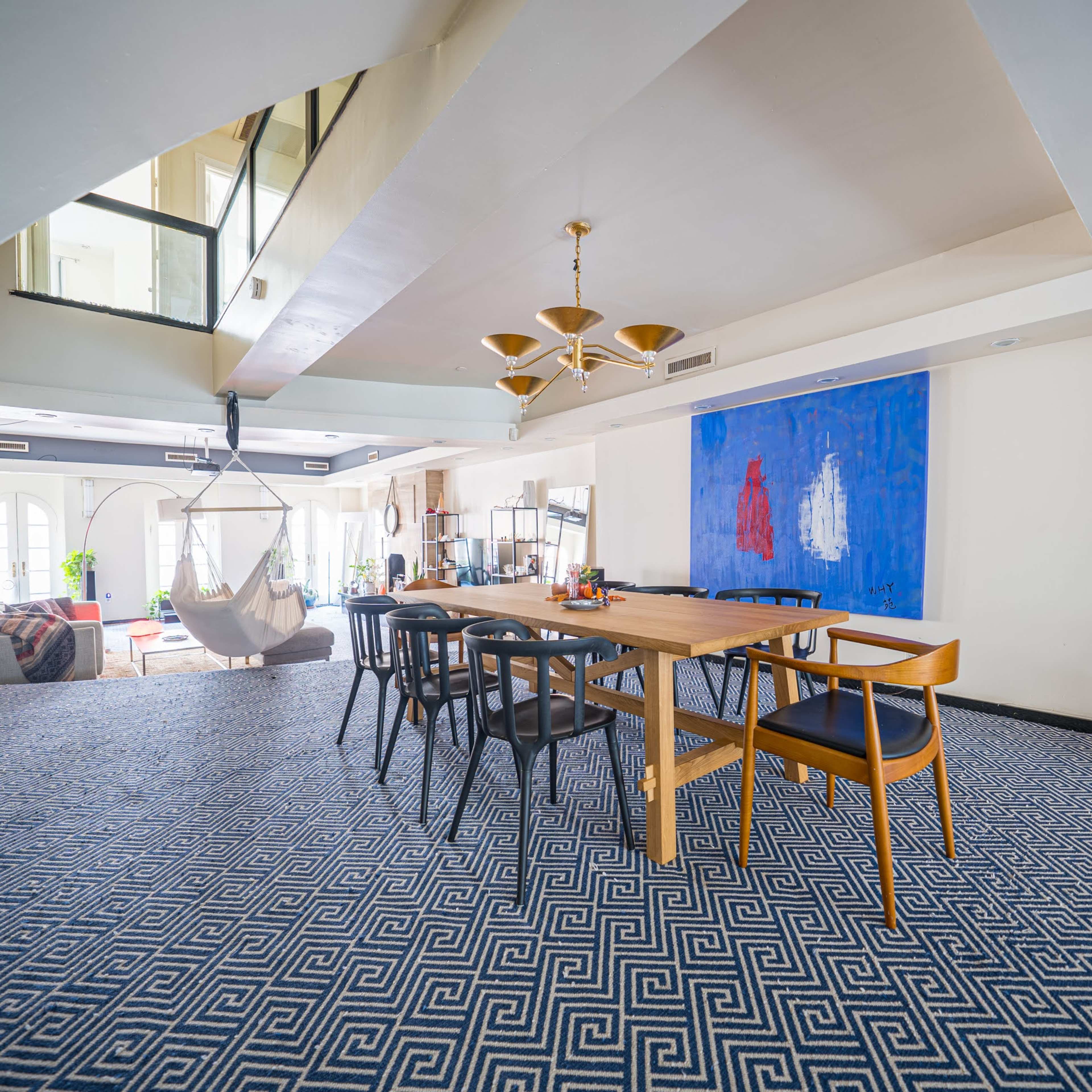 A spacious indoor area with a large dining table surrounded by chairs, a hammock hanging nearby, and a prominent blue artwork on the wall.