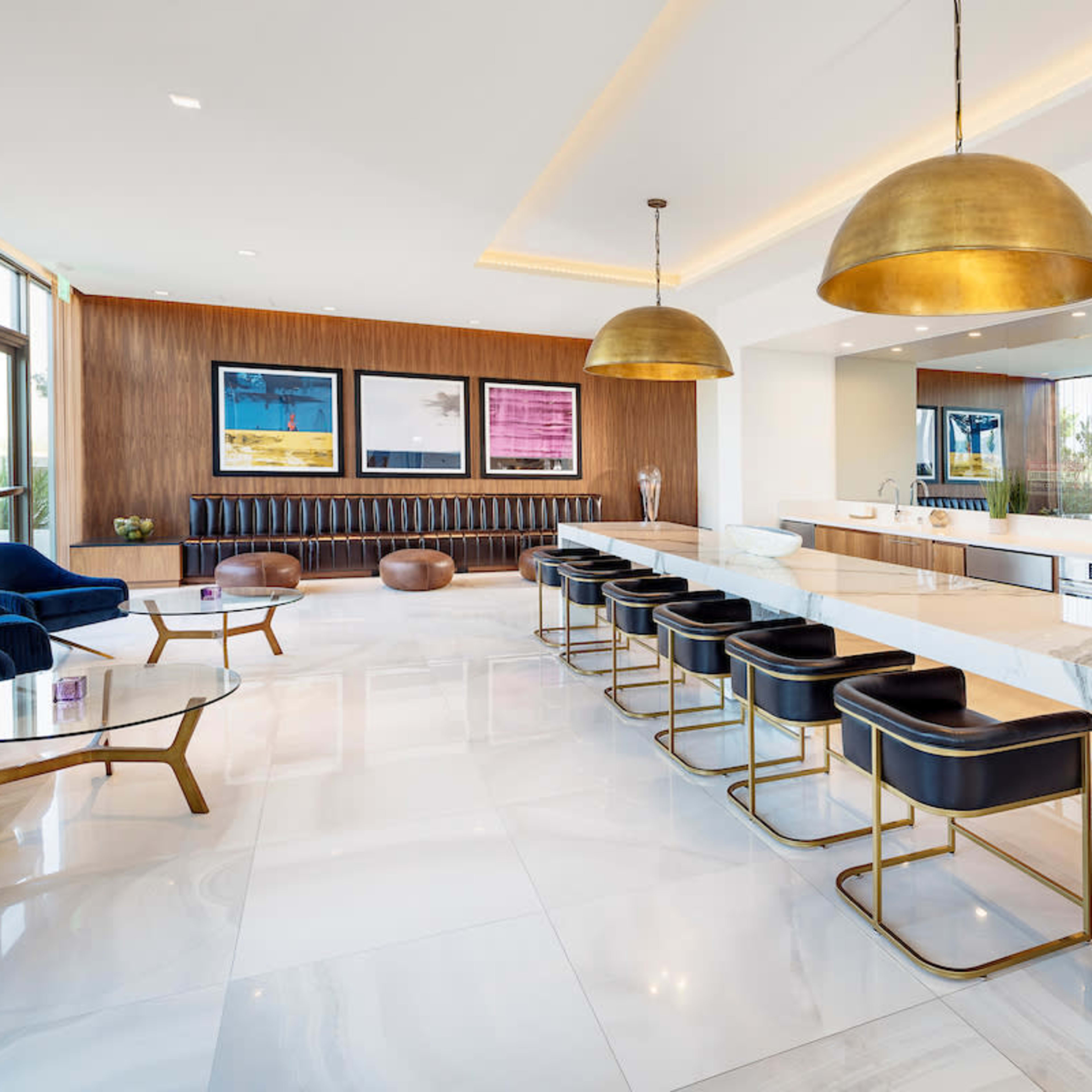 A modern kitchen area features a marble countertop bar with gold pendant lights and black stools, surrounded by sleek furniture and large windows.