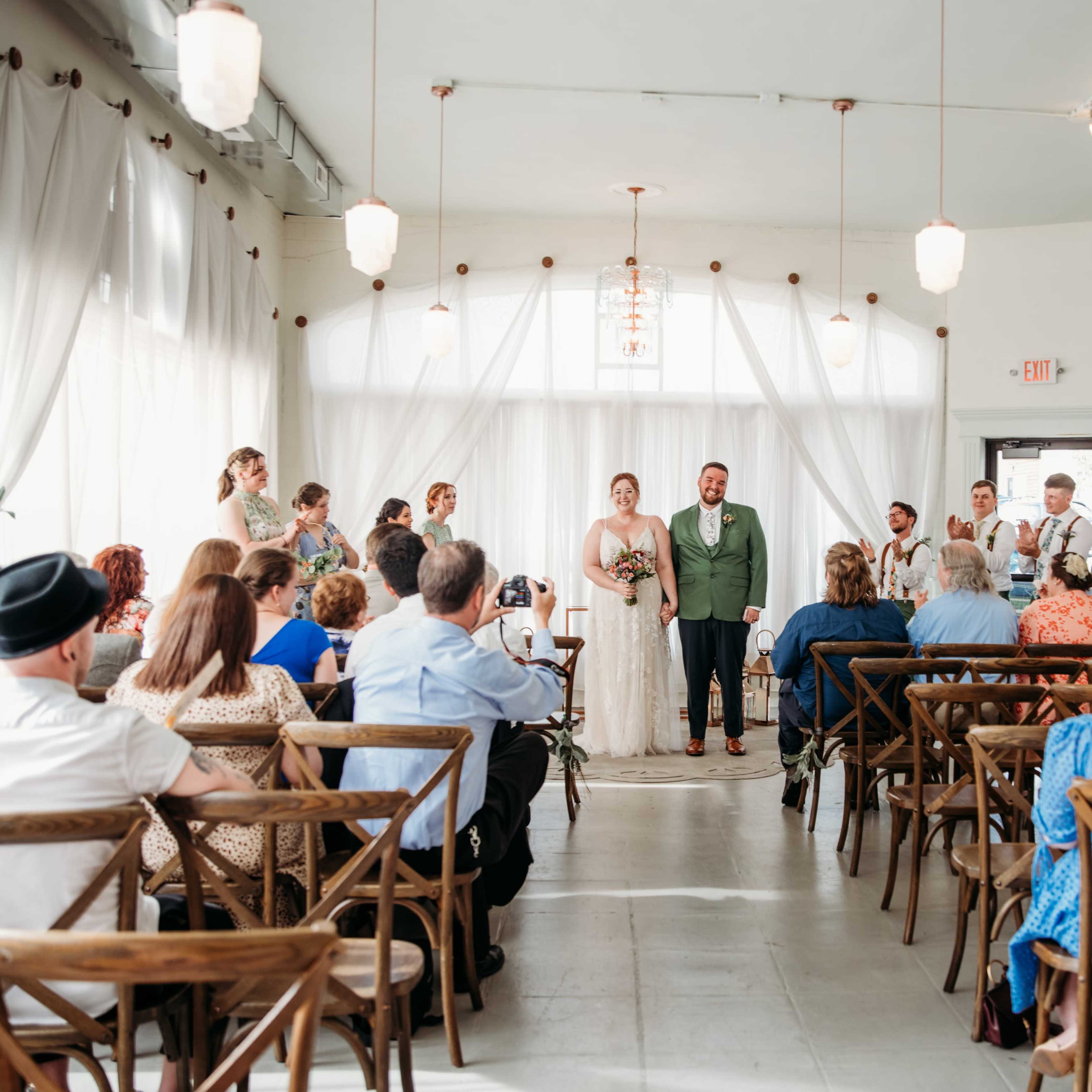 A wedding ceremony is taking place in a well-lit venue, with guests seated on wooden chairs and the bridal party standing at the front.