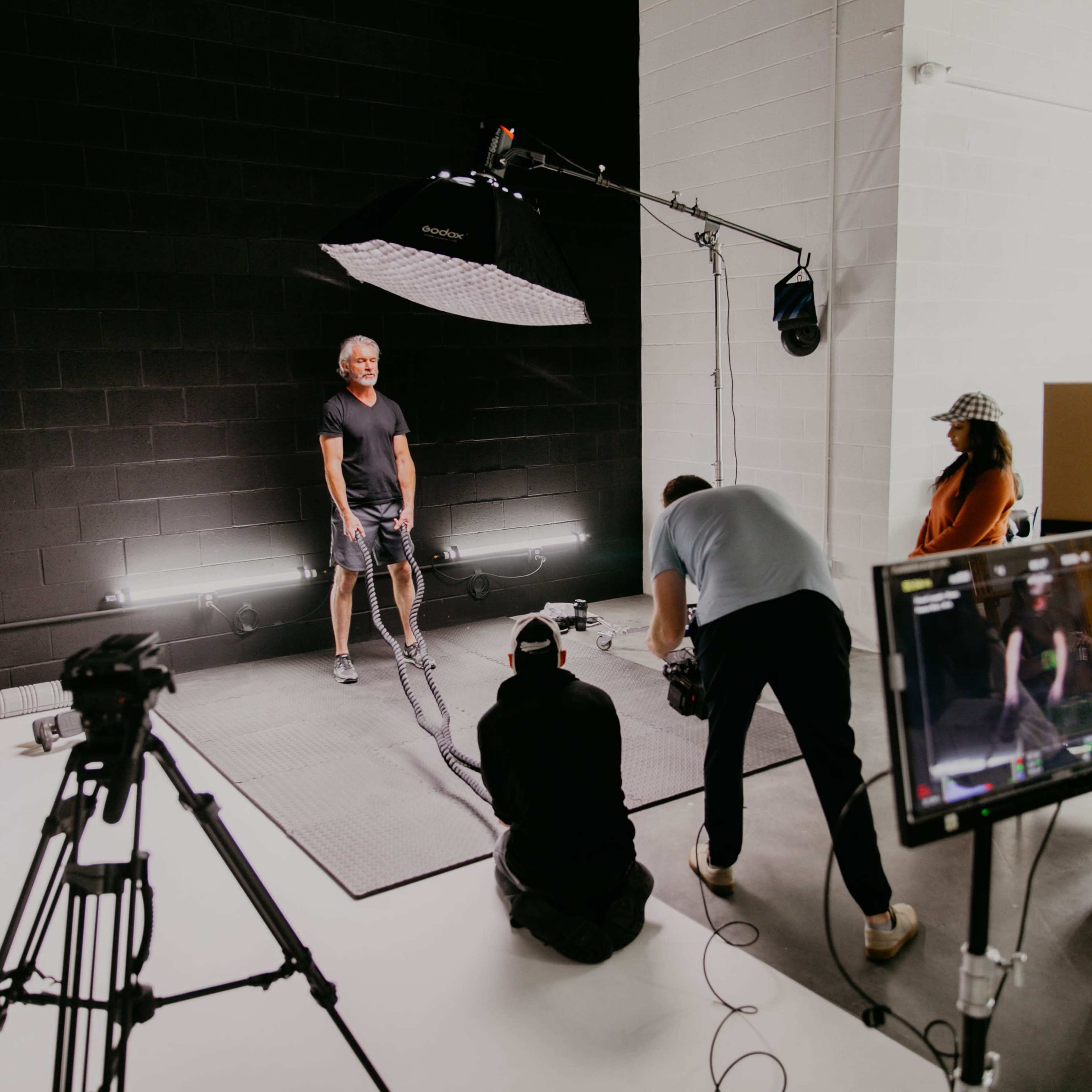 A photoshoot is taking place in a studio with a subject standing on a mat under a large light source while crew members manage the setup and monitor the scene.