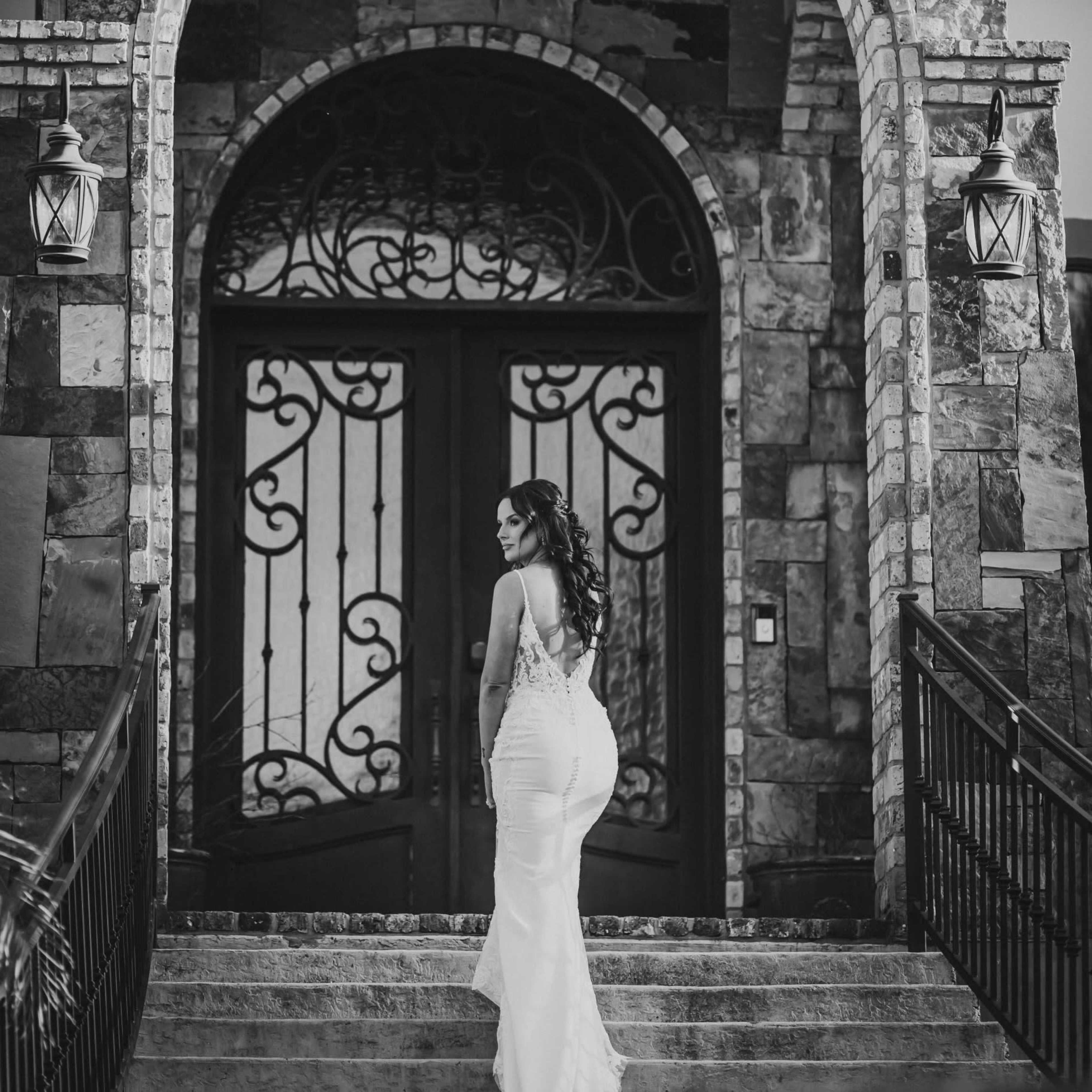 A woman in a white dress stands on steps leading to a grand stone entrance with intricate doors.