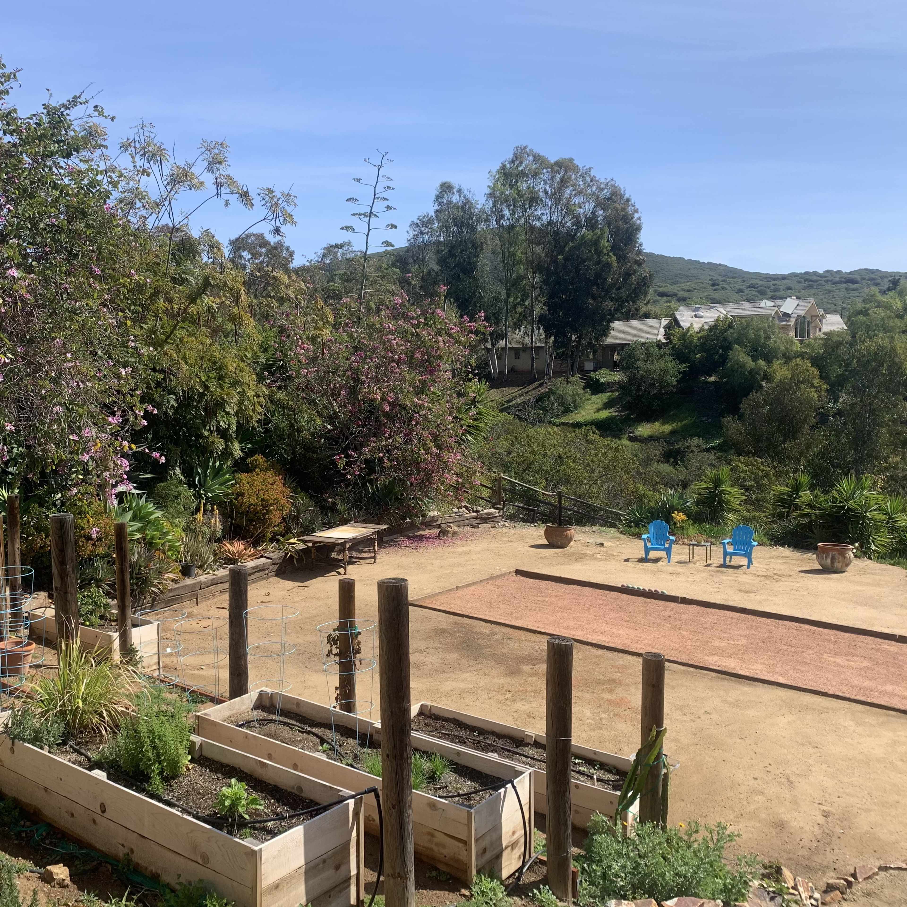 A landscaped garden area with raised planter boxes, a dirt patch, and blue chairs set against a backdrop of trees and hills.