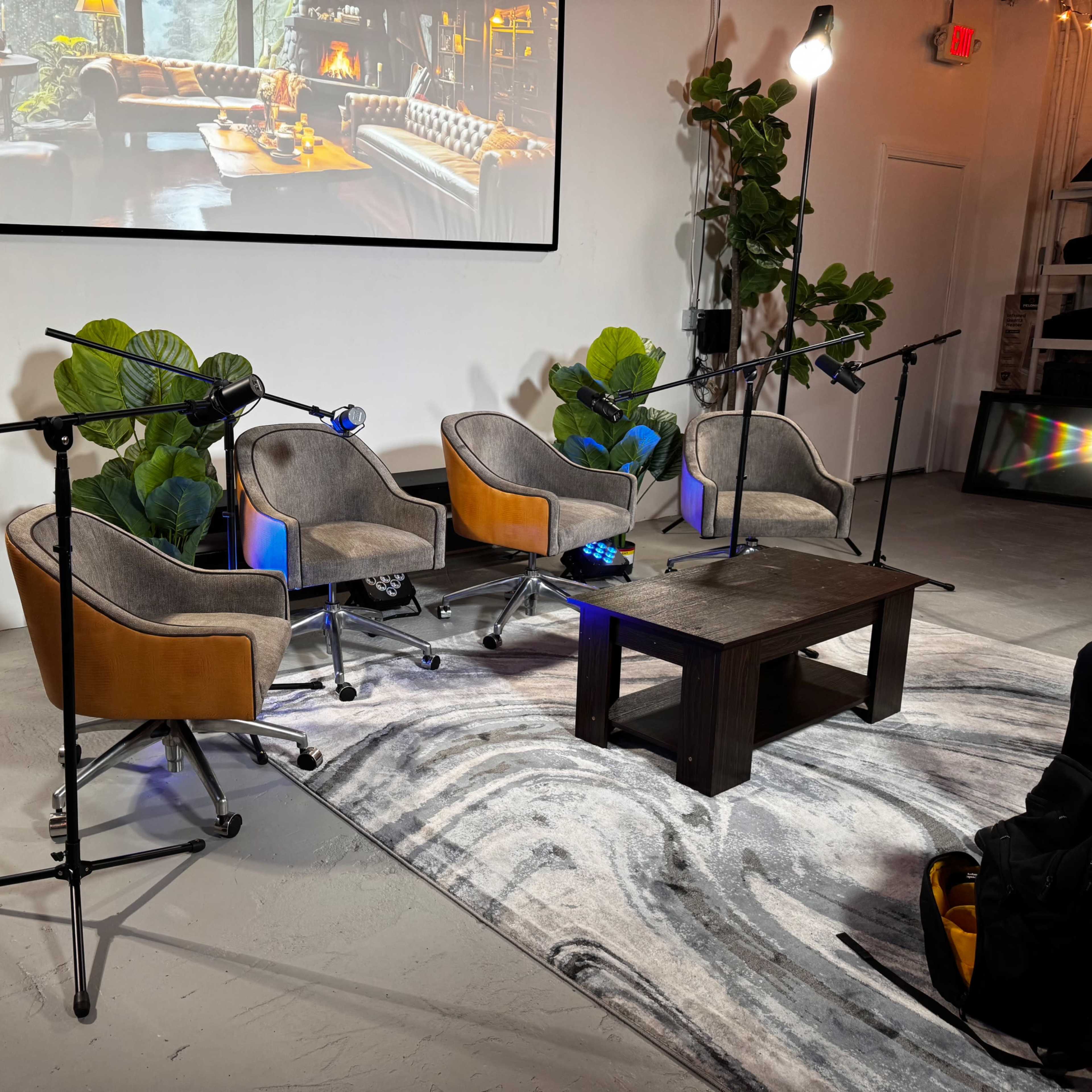 A cozy event space with four upholstered chairs arranged around a low wooden table, equipped with microphones and surrounded by large green plants.