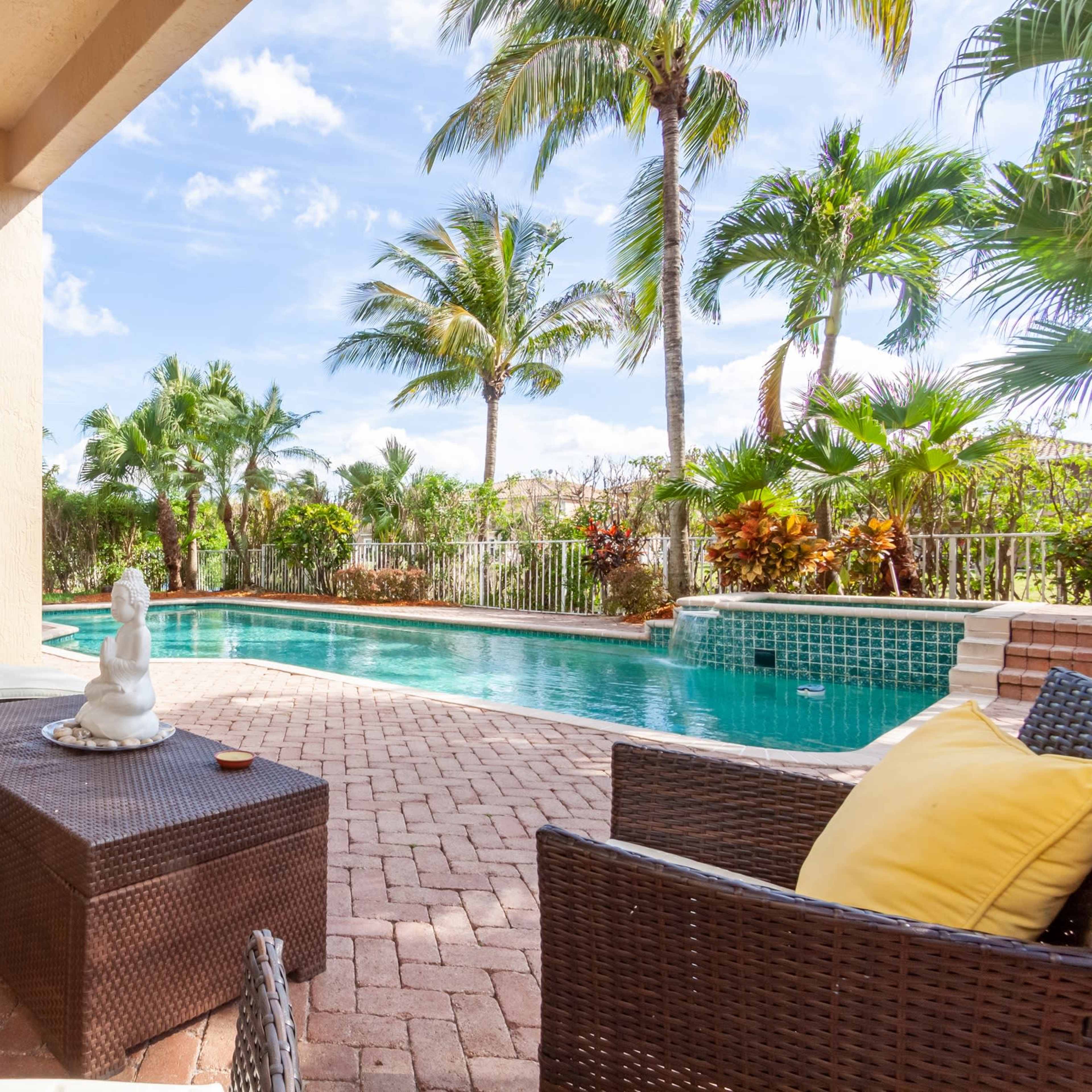 A patio area with a pool surrounded by palm trees and tropical foliage, featuring lounge chairs and a coffee table.