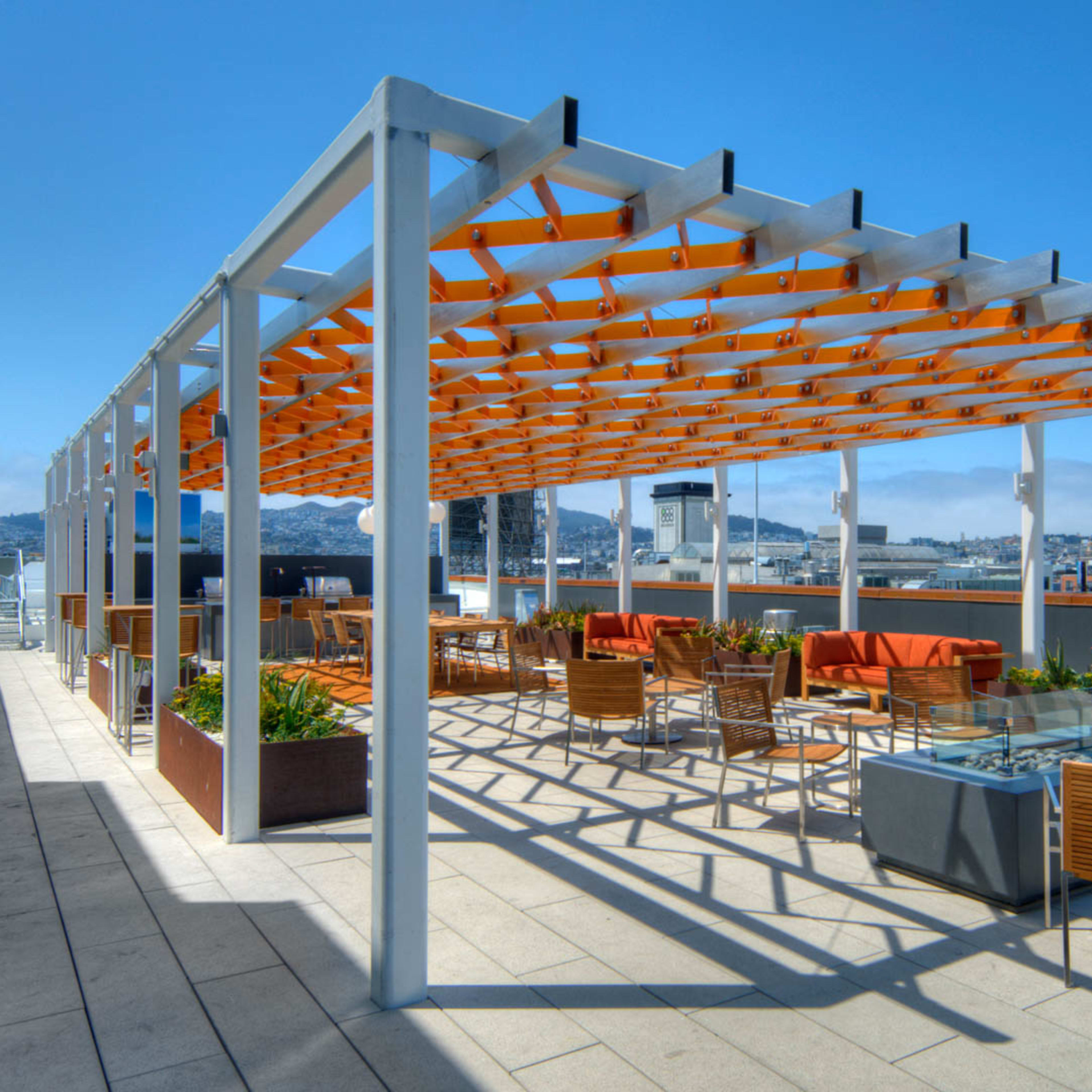 A rooftop terrace features a pergola with orange accents, surrounded by tables and chairs, offering a view of city buildings and a clear sky.