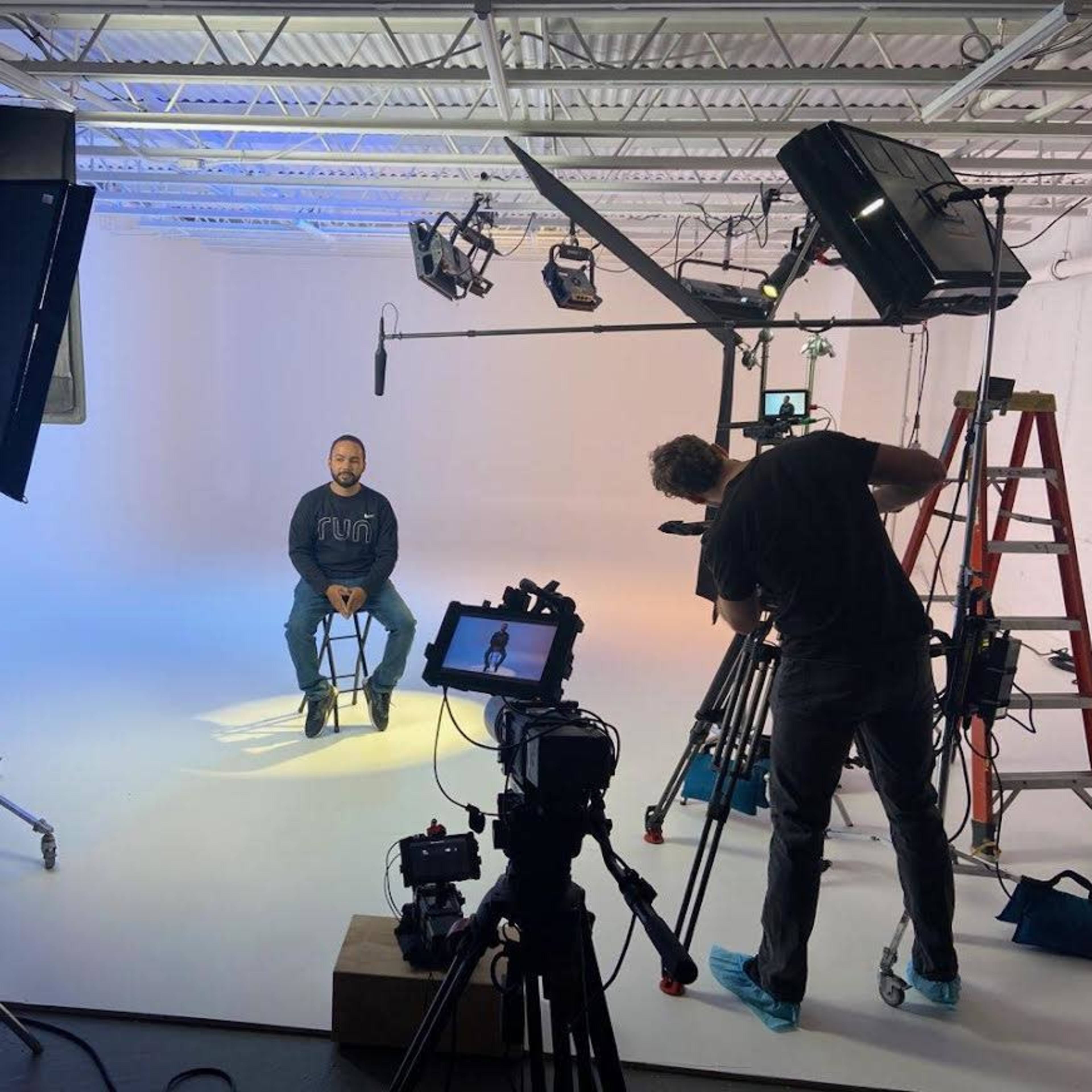 A person sits on a stool in a well-lit studio with cameras and lighting equipment set up around them.