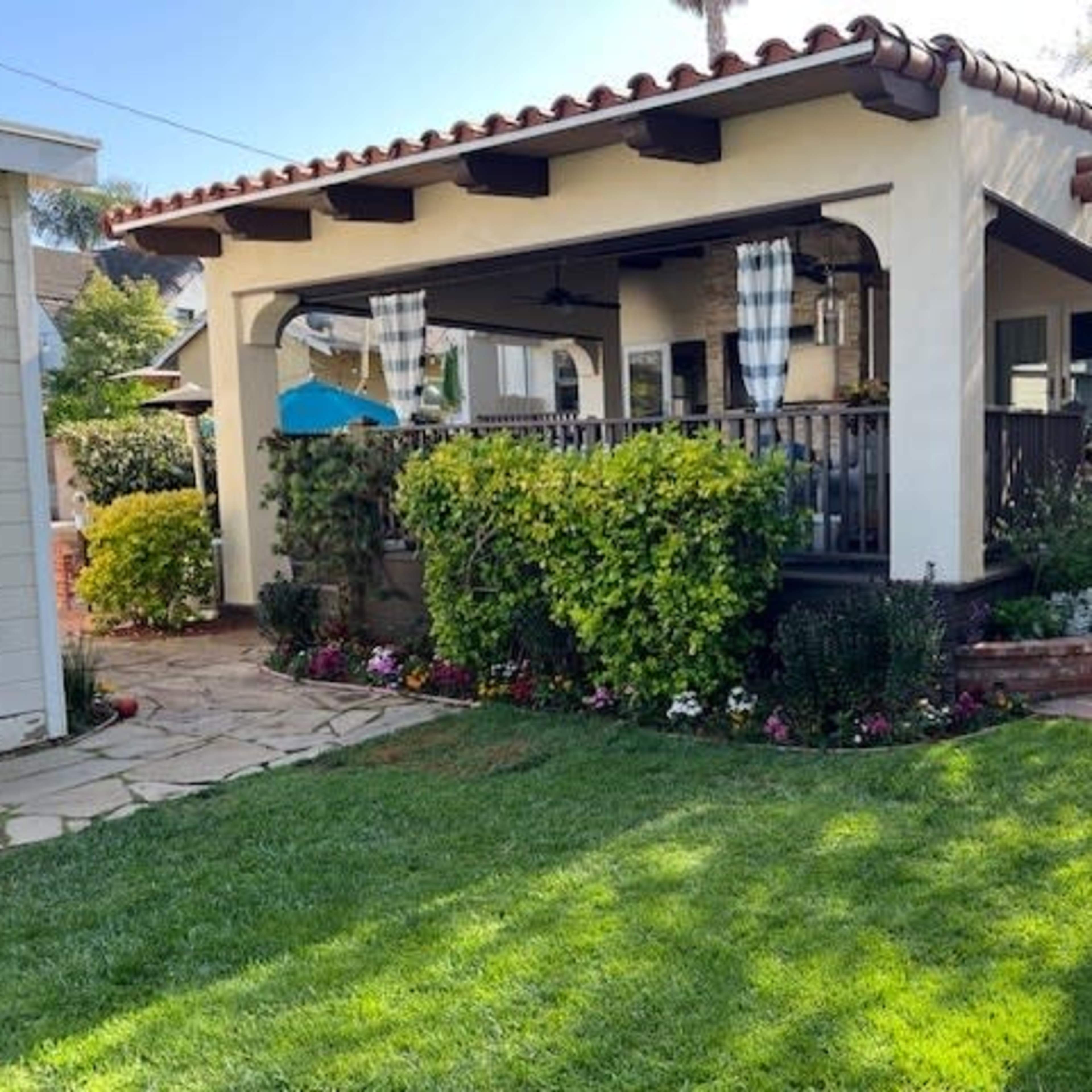 A well-maintained, single-story house features a covered porch surrounded by lush greenery and vibrant flowers in the front yard.