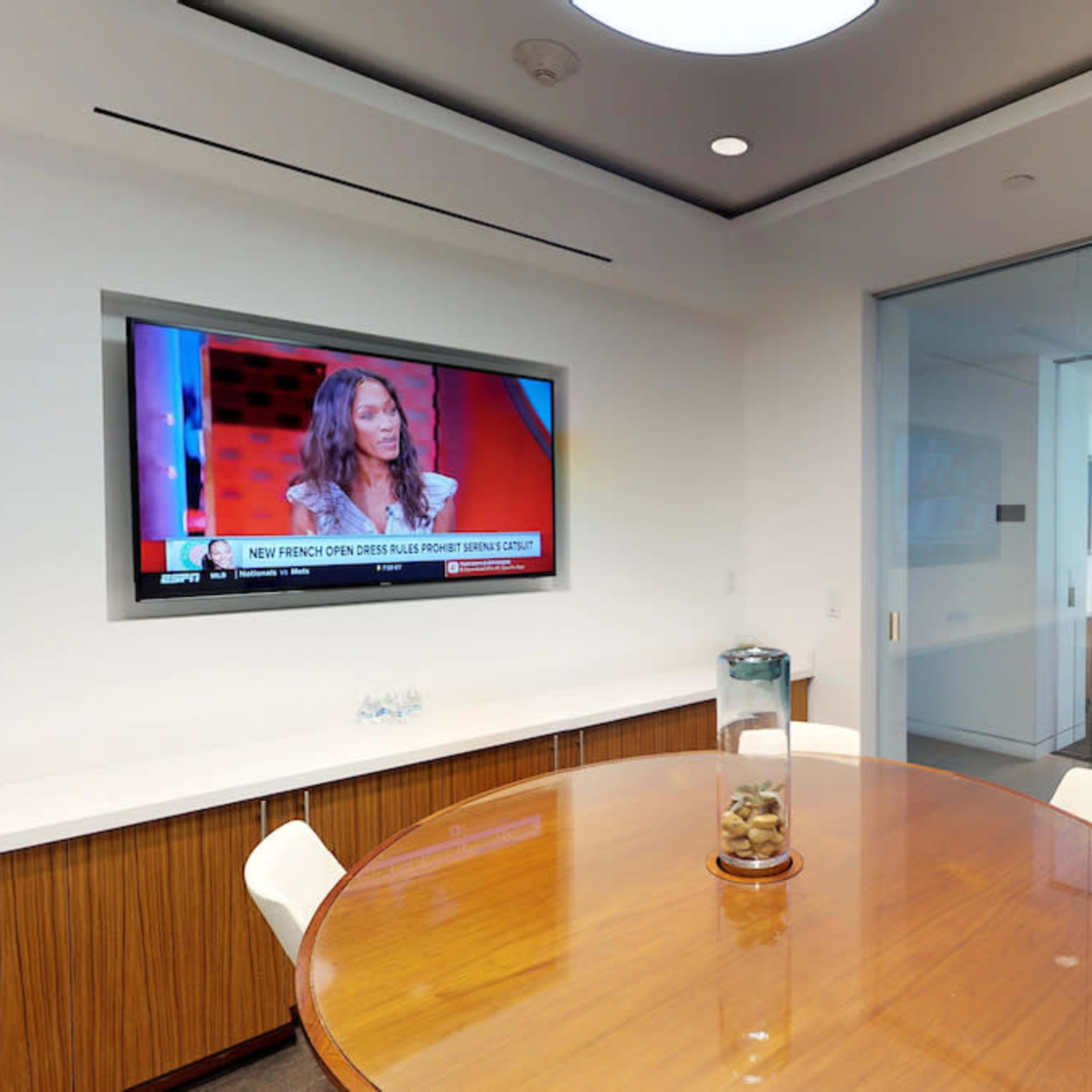 A conference room features a round table with white chairs and a television displaying a news program on the wall.
