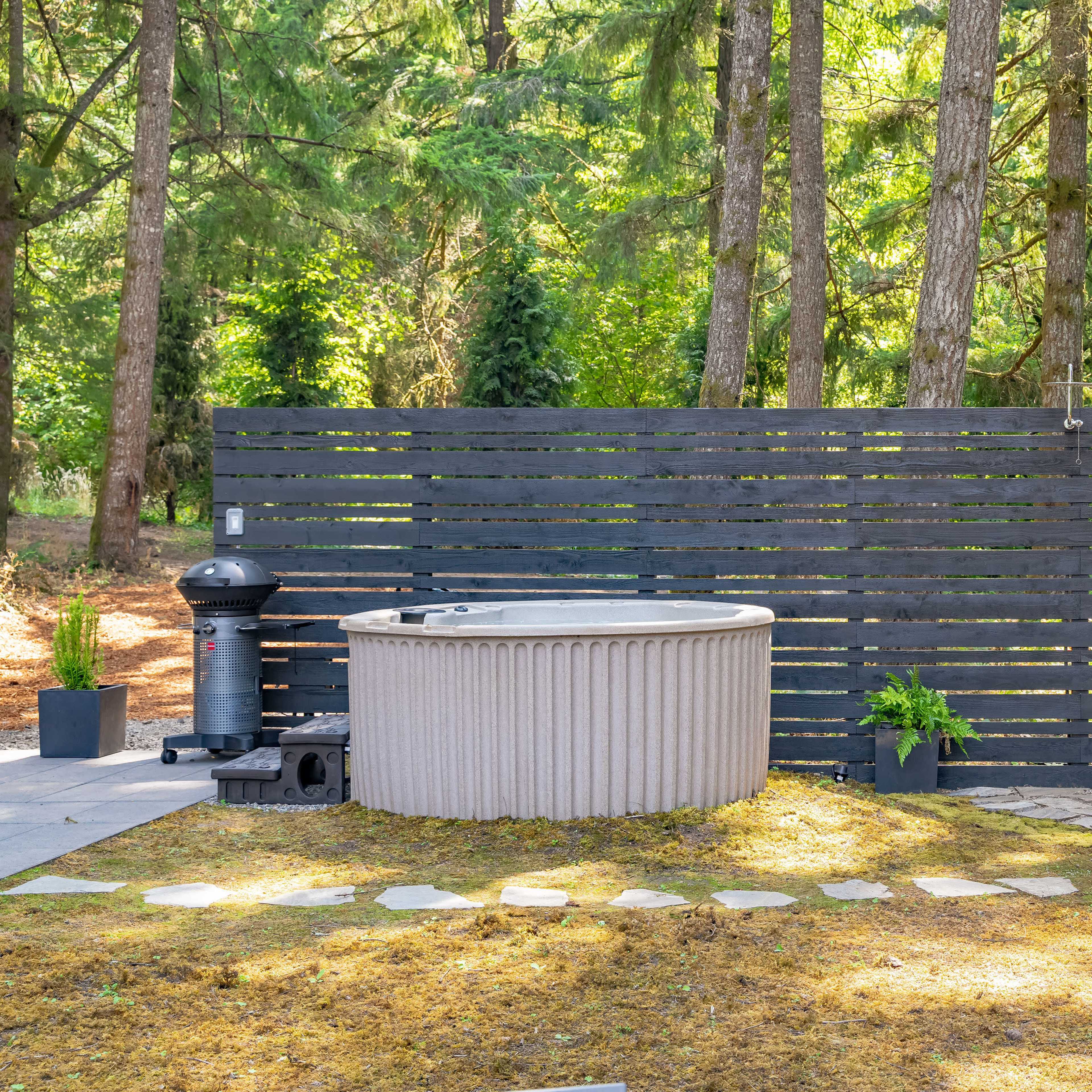 A landscaped outdoor area featuring a hot tub, a barrel sauna, and a wooden privacy screen, surrounded by trees and stone pathways.