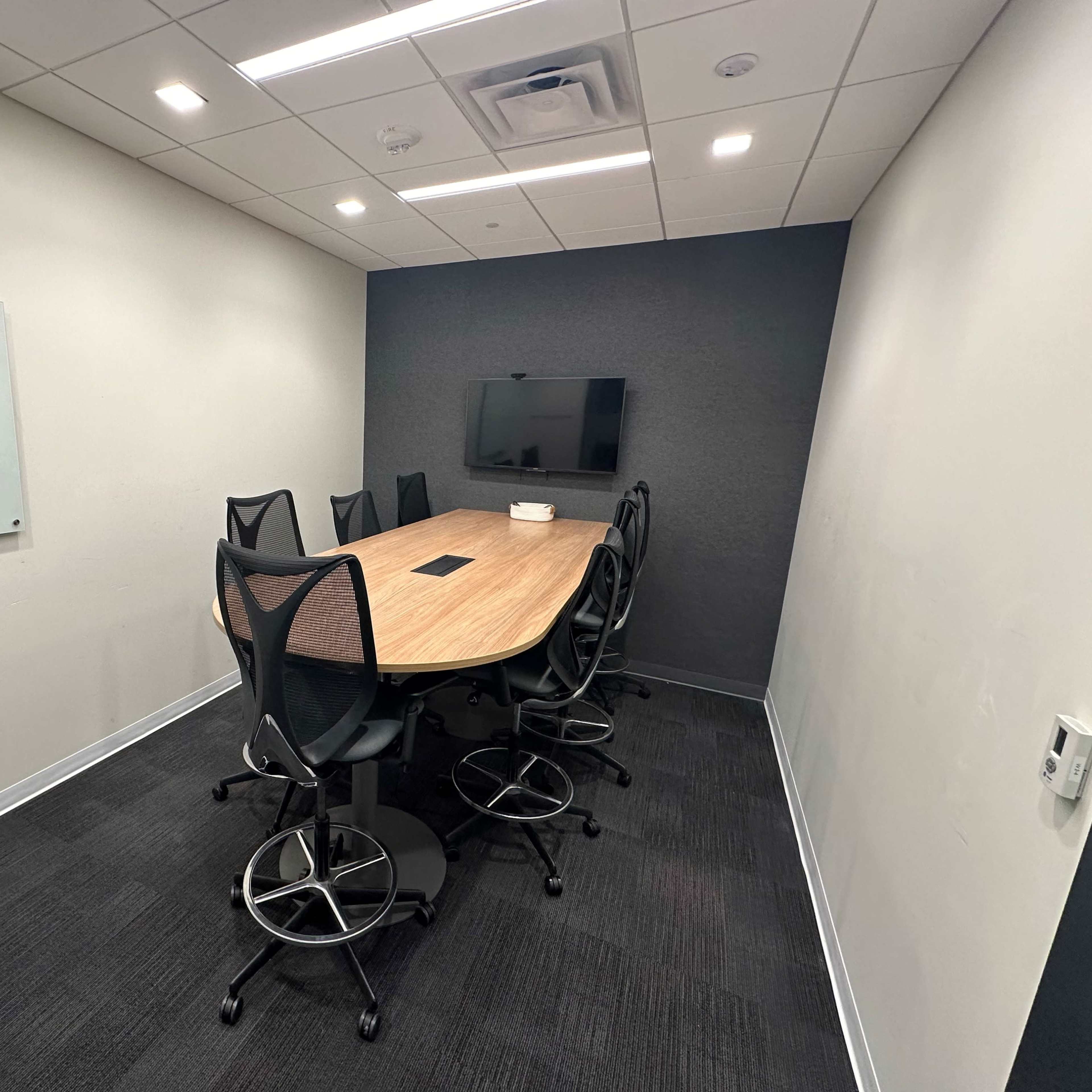 A meeting room features a large wooden table surrounded by six black ergonomic chairs, a wall-mounted TV, and a whiteboard.