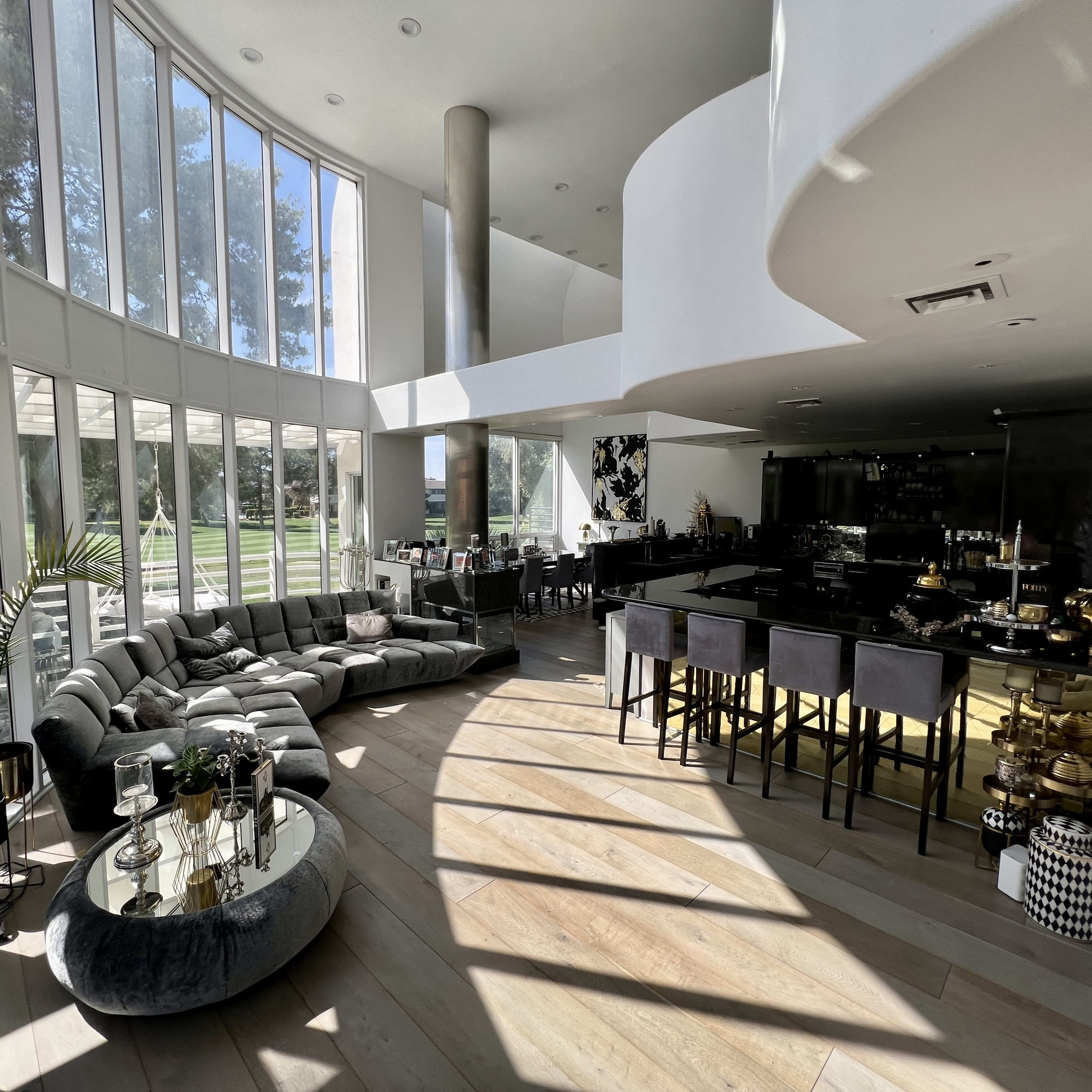 The spacious interior features a sectional sofa and a modern kitchen, illuminated by large windows that overlook a green landscape.