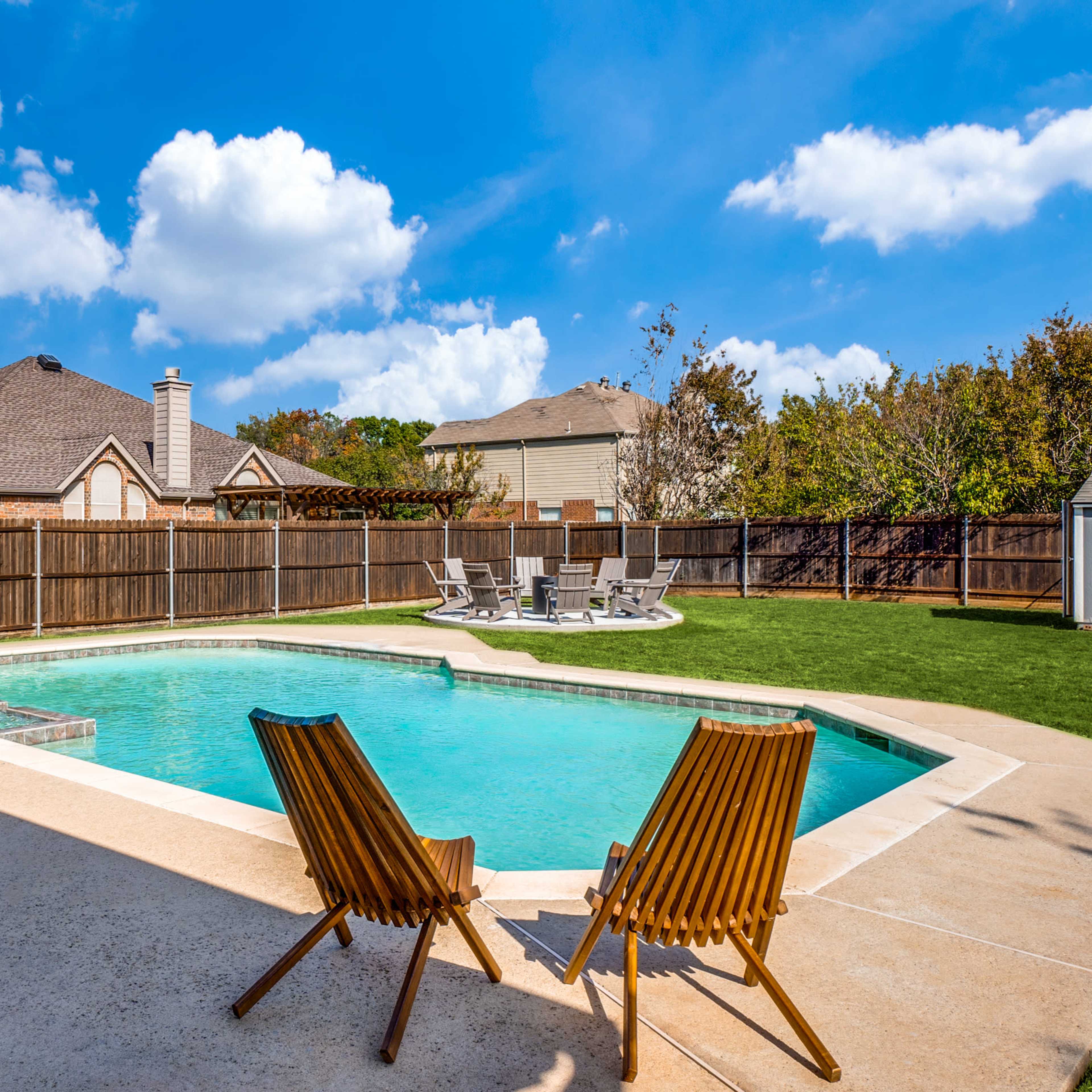 A backyard with a swimming pool, two wooden lounge chairs, and a fenced area with houses in the background.