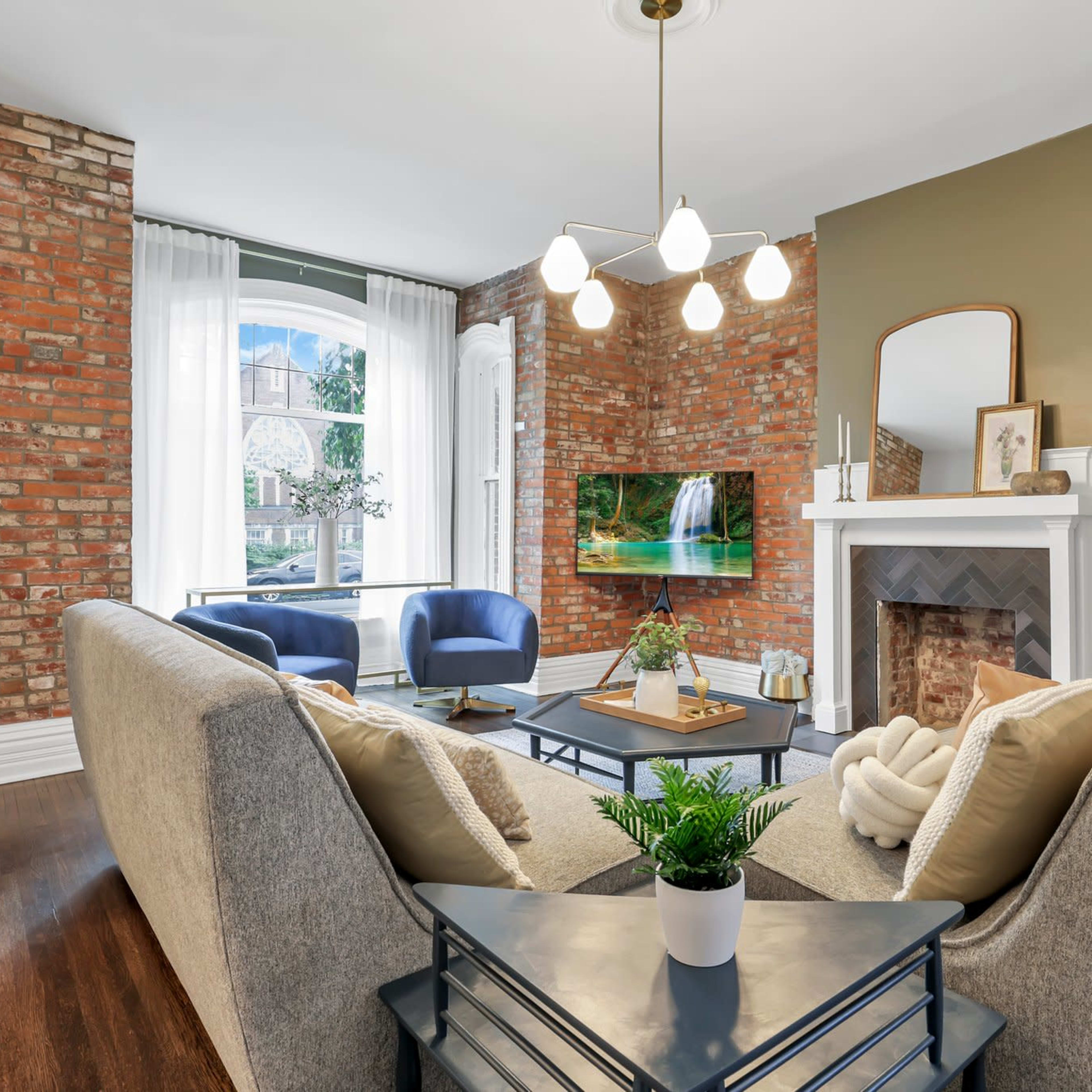 A cozy living room features a gray sofa, a coffee table, and a fireplace, with exposed brick walls and a large window displaying a view of the outdoors.