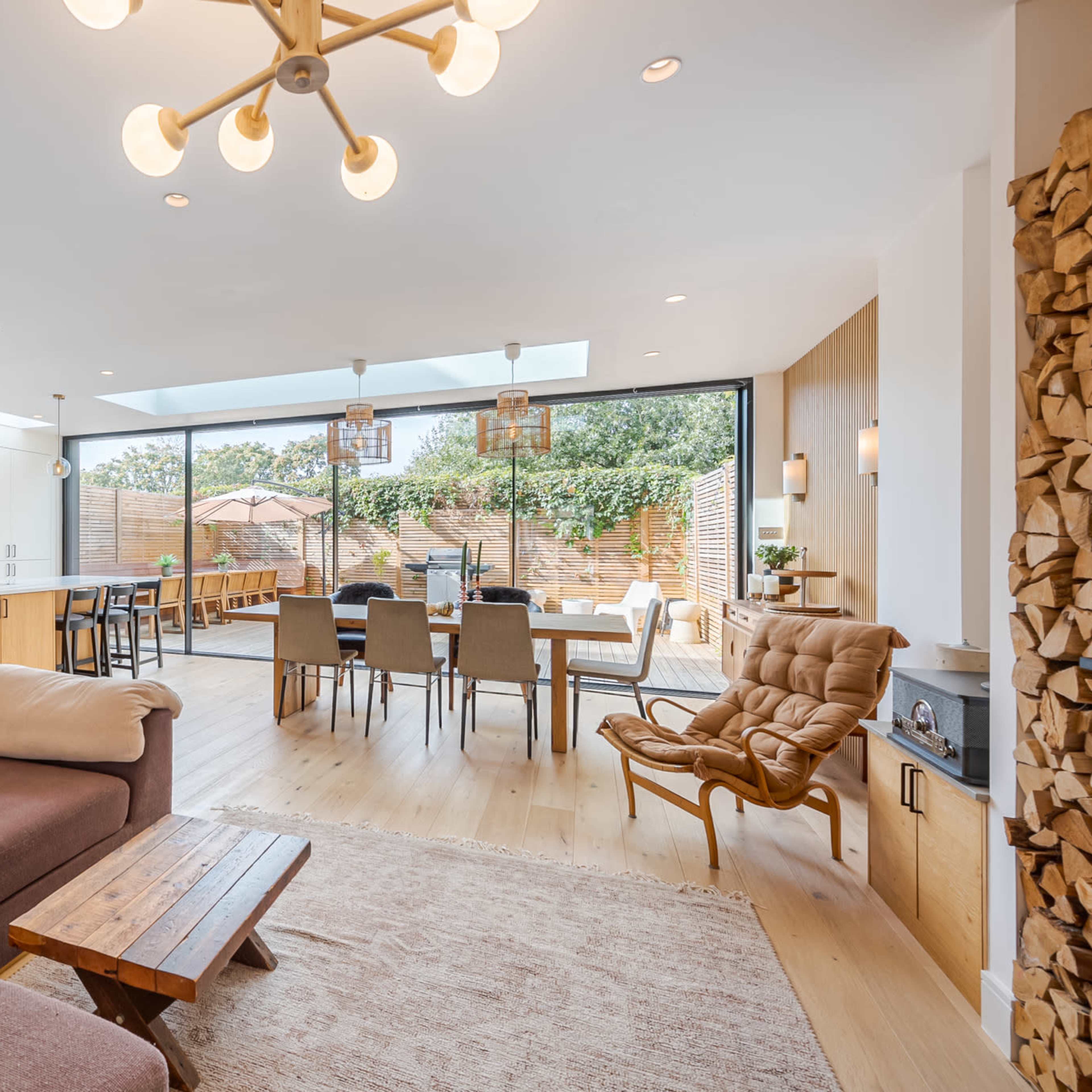A modern living and dining area with large windows, featuring a kitchen on one side, a dining table in the center, and a cozy seating arrangement with a rug in front.