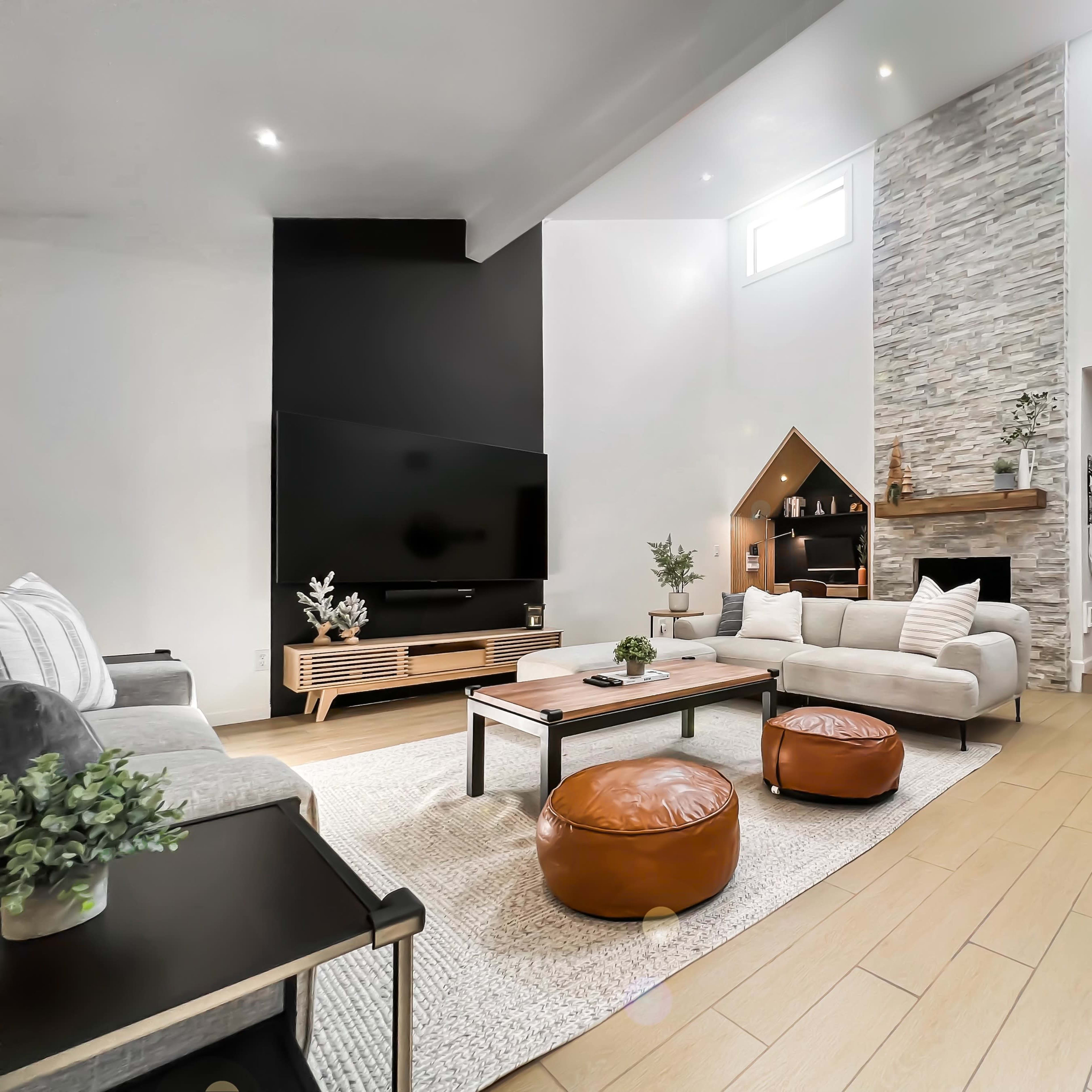 A modern living room featuring a large flat-screen TV, a stone fireplace, and a mix of seating including a sectional sofa and leather ottomans.