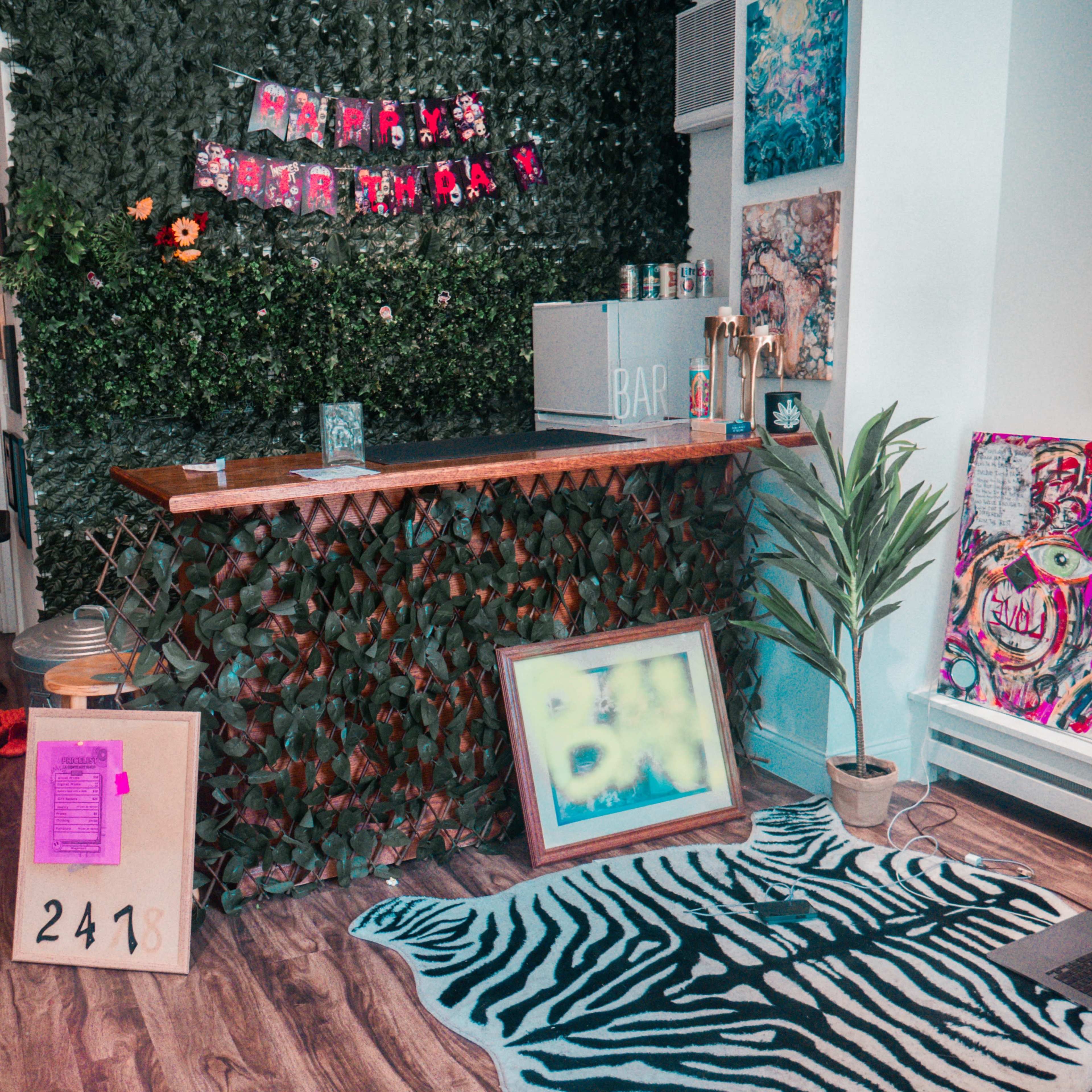 A small reception area with a wooden bar adorned with greenery, a framed picture on the floor, and colorful artwork on the walls.