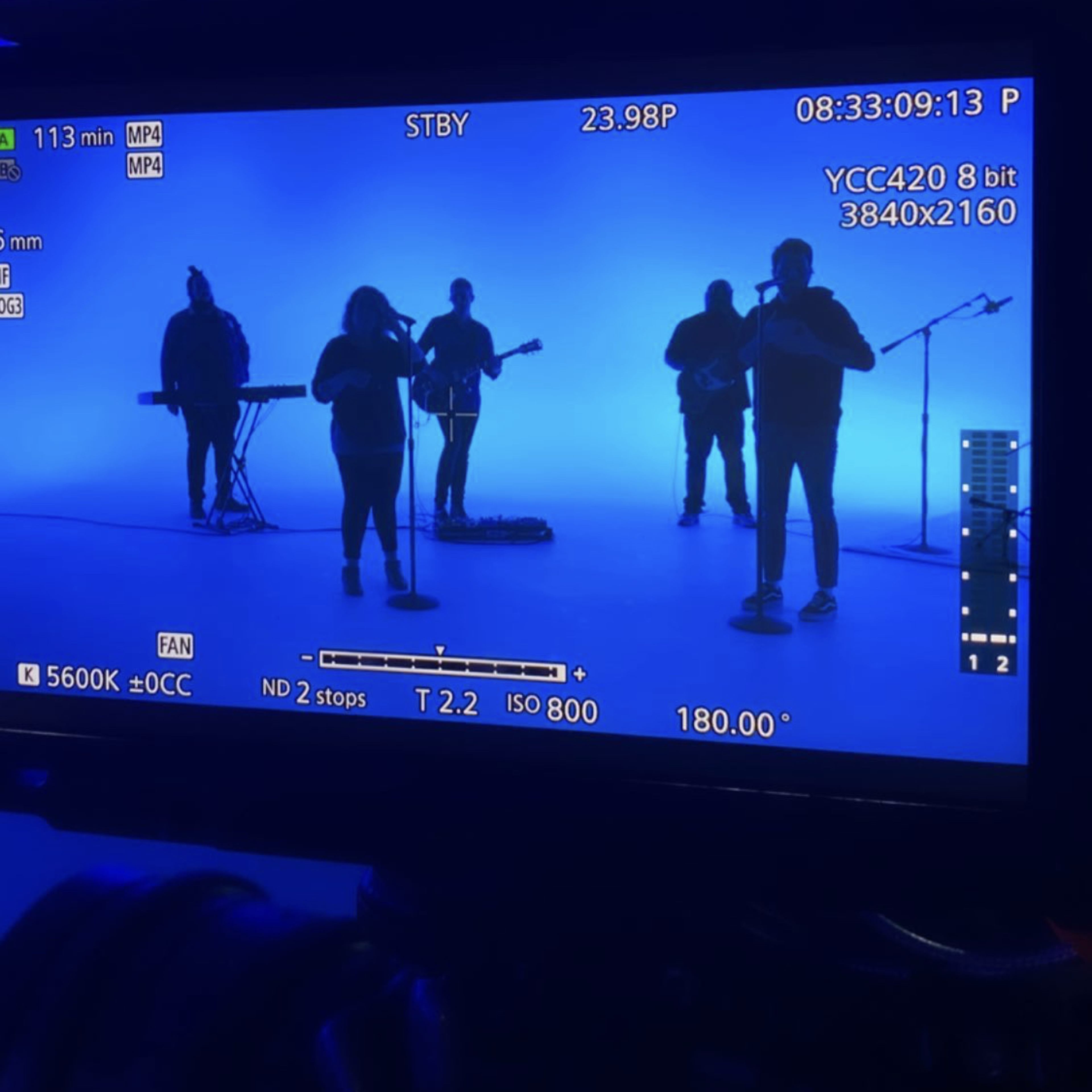 A band performs under blue lighting, with a camera recording the scene from the foreground.