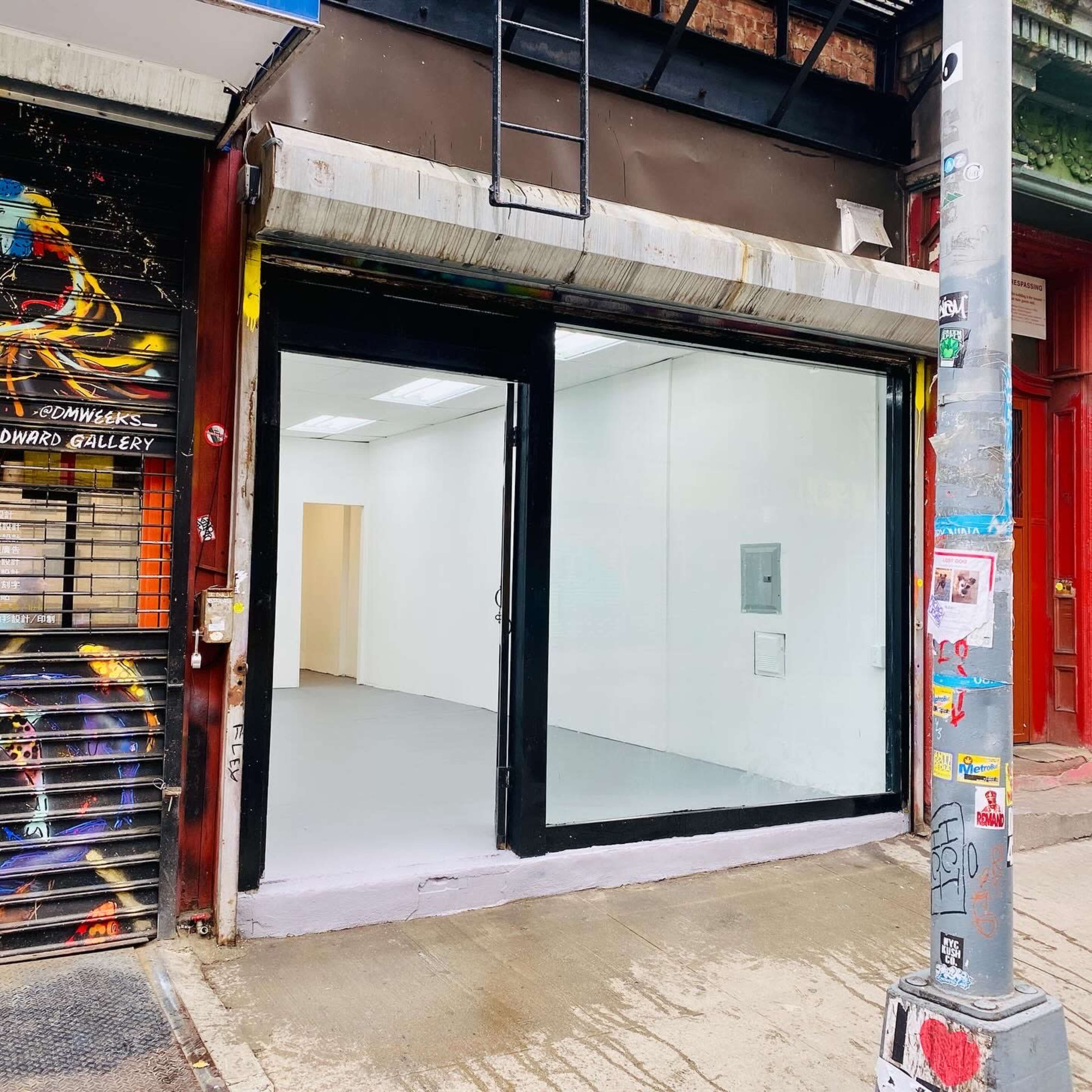 A storefront with large glass windows and a blank interior, located on a city street featuring colorful street art and a utility pole adorned with various stickers.