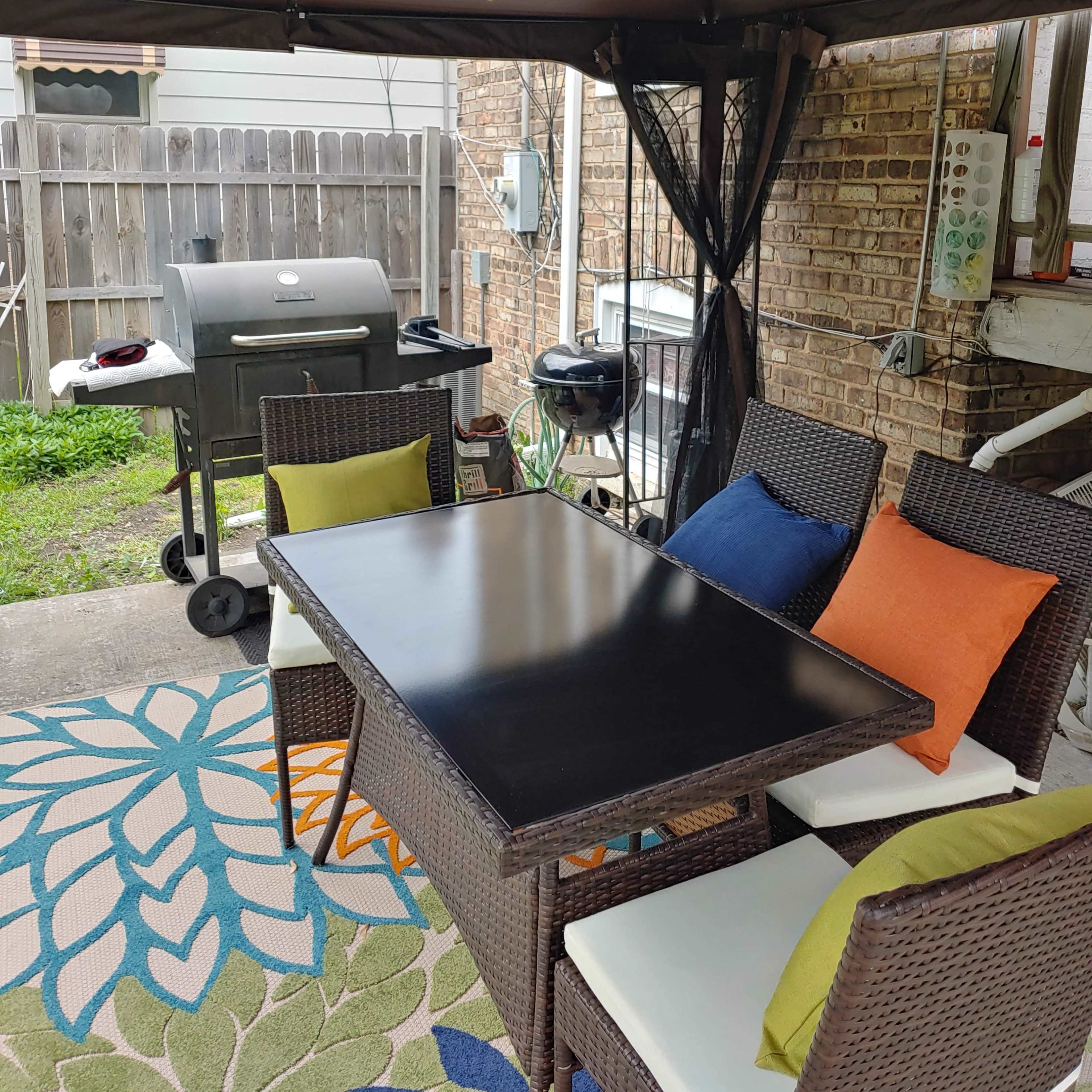 A patio area features a black table surrounded by four chairs, with a grill positioned nearby under a gazebo.
