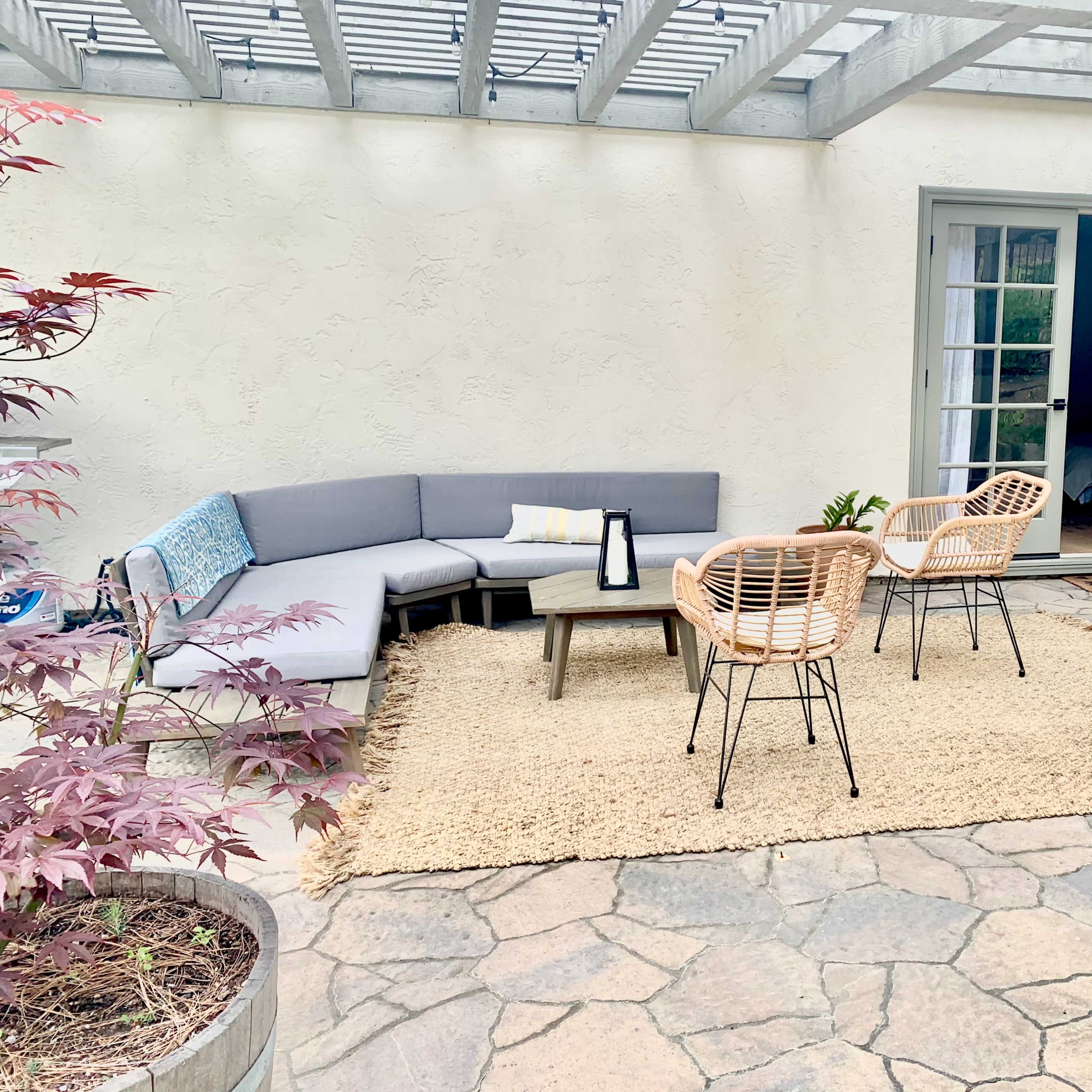 A patio features a sectional sofa, two chairs, and a coffee table on a stone surface, with a door leading to an interior room.