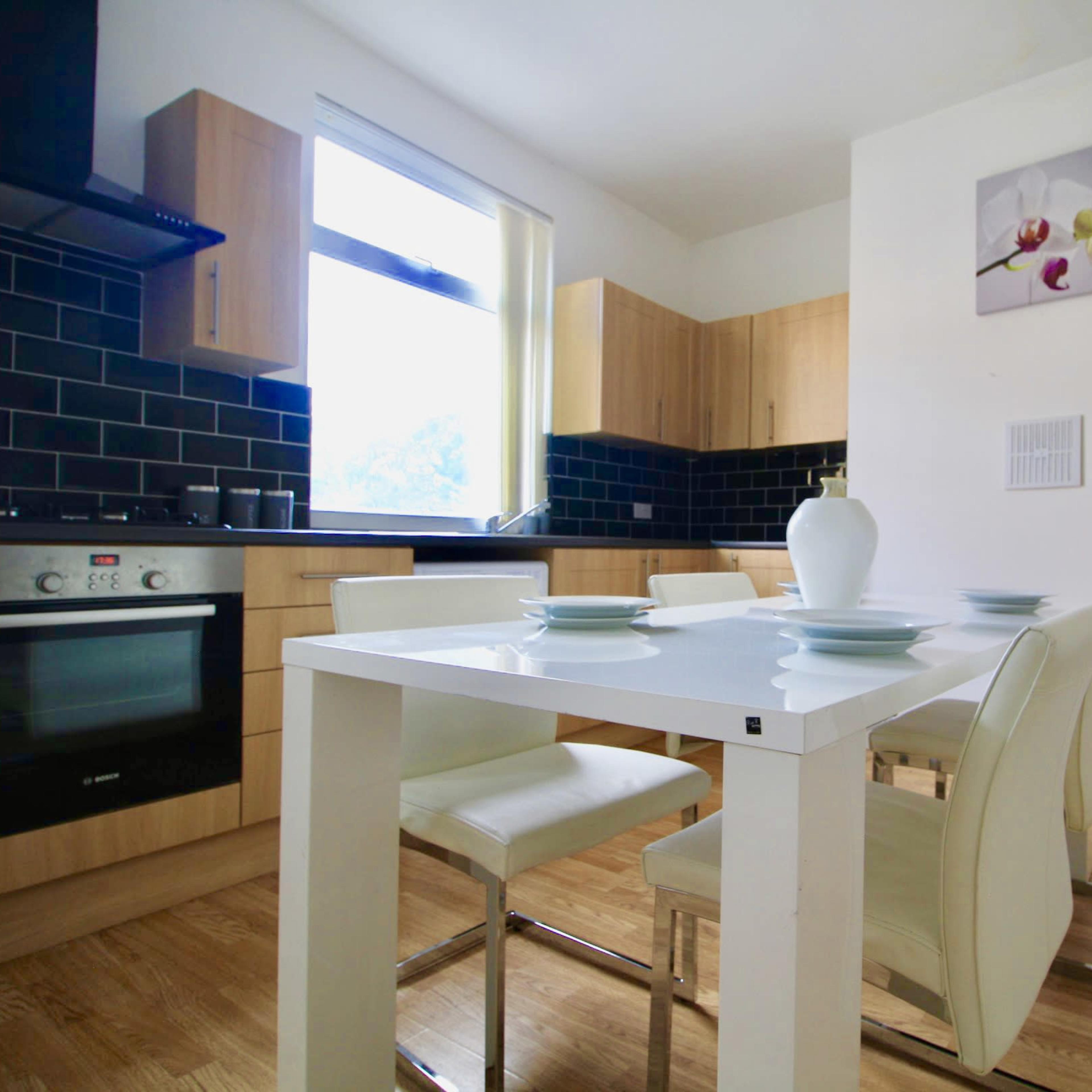 A modern kitchen features wooden cabinetry, black tiled walls, and a white dining table with four chairs.