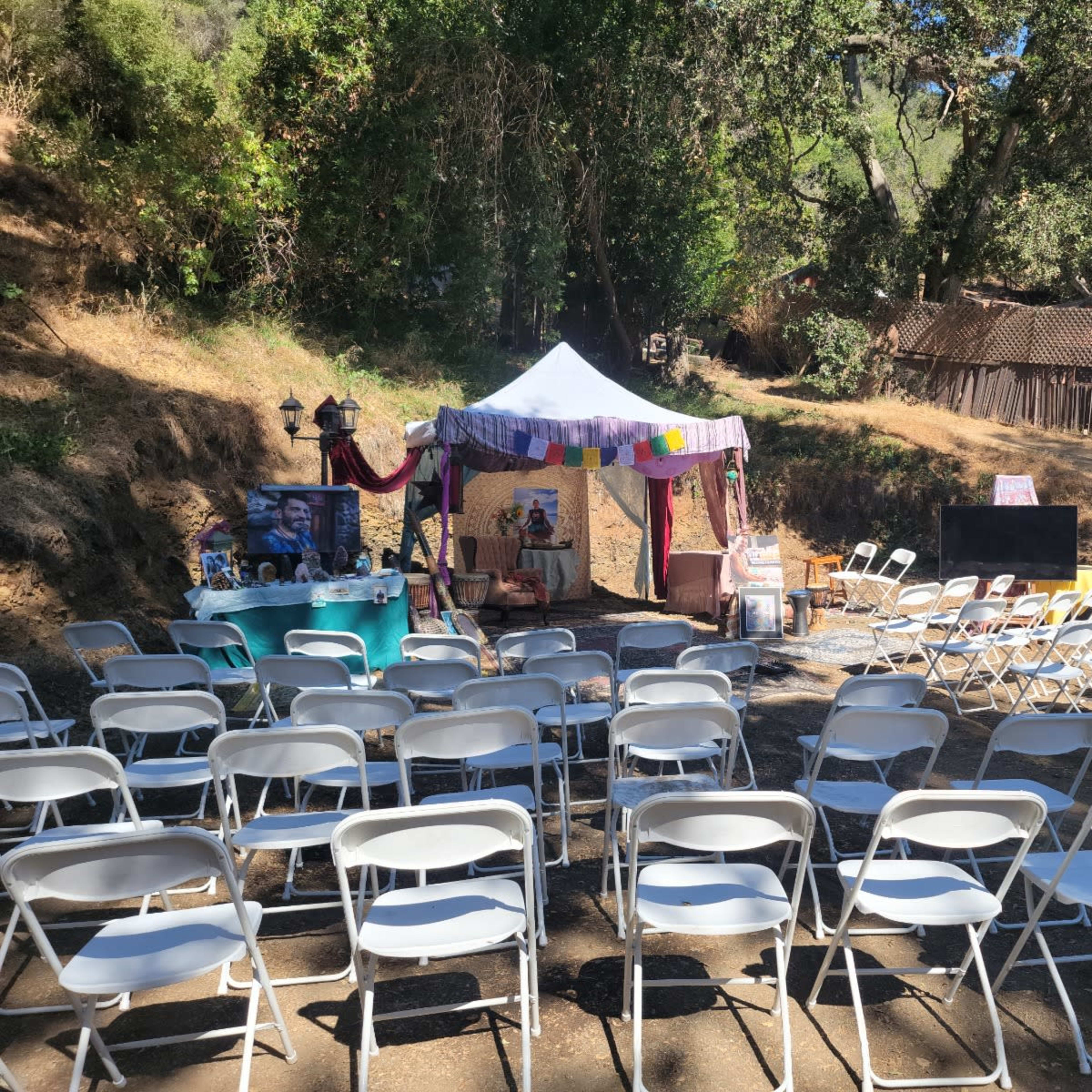 A tent with colorful decorations is set up in a clearing, facing rows of white folding chairs, with a rustic building visible in the background.