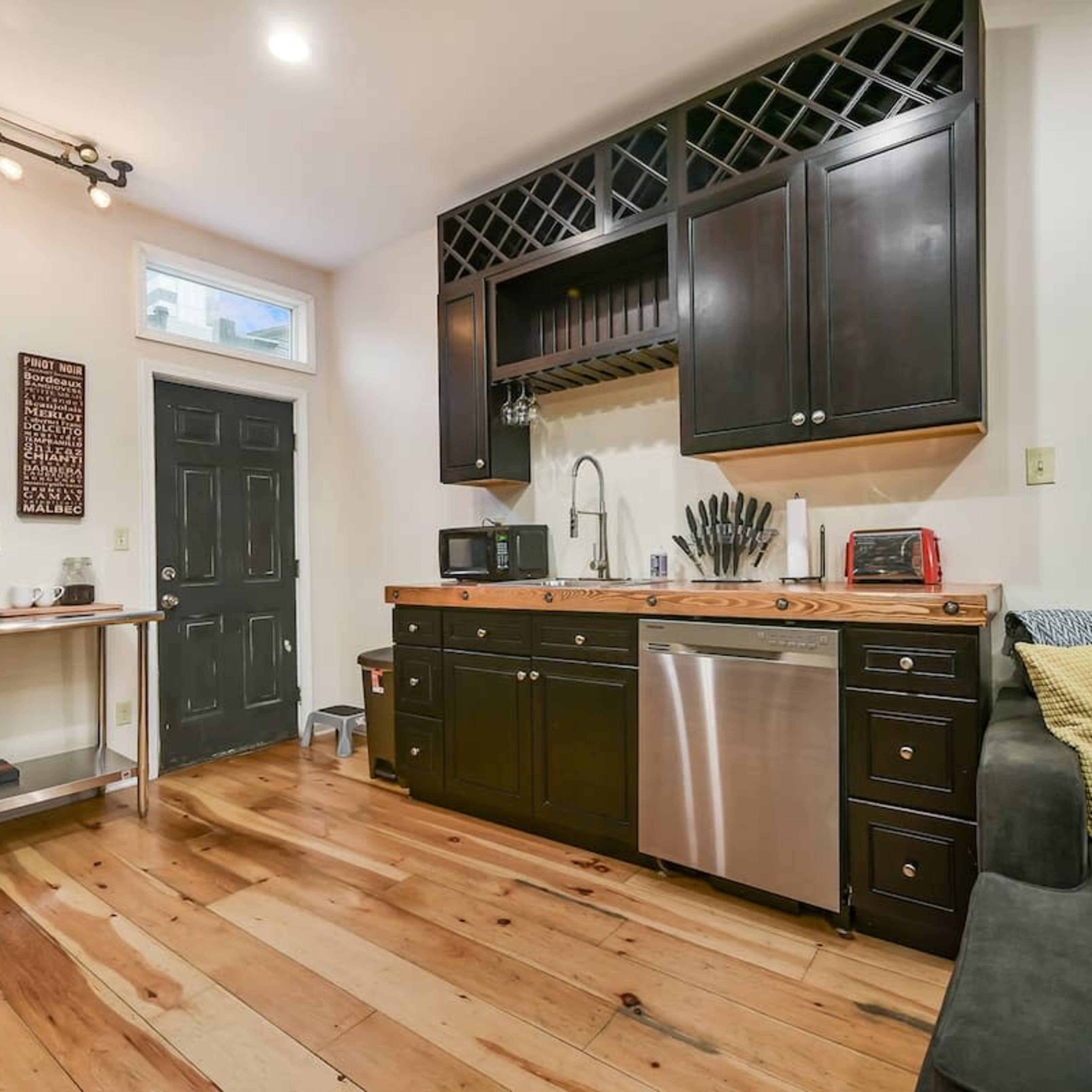 A cozy kitchenette with dark cabinets, a stainless steel sink, and wooden countertops, adjacent to a living area with light wooden flooring.