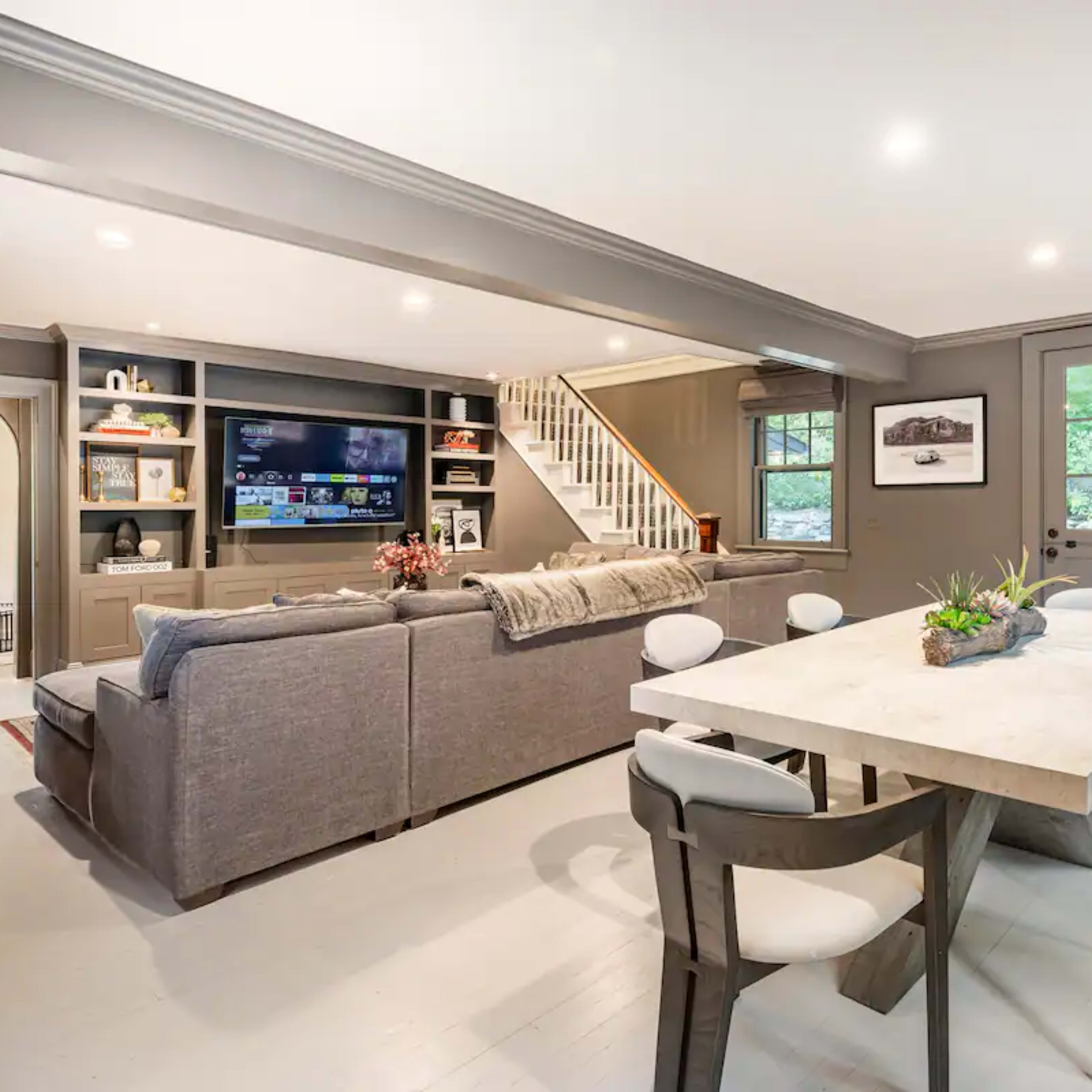 A spacious living area features a gray sofa, a large television on a built-in shelf, and a dining table surrounded by modern chairs.