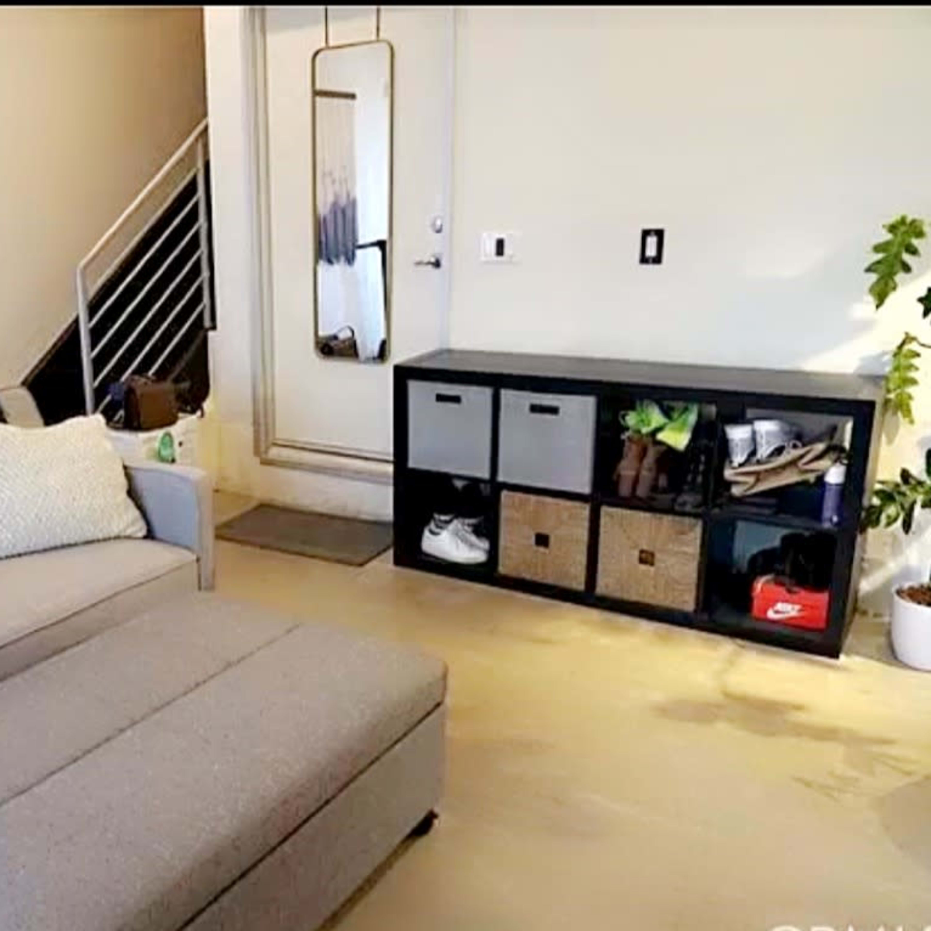 A modern living room featuring a gray couch with decorative pillows, an ottoman, and a storage unit with baskets and shoes against a wall.