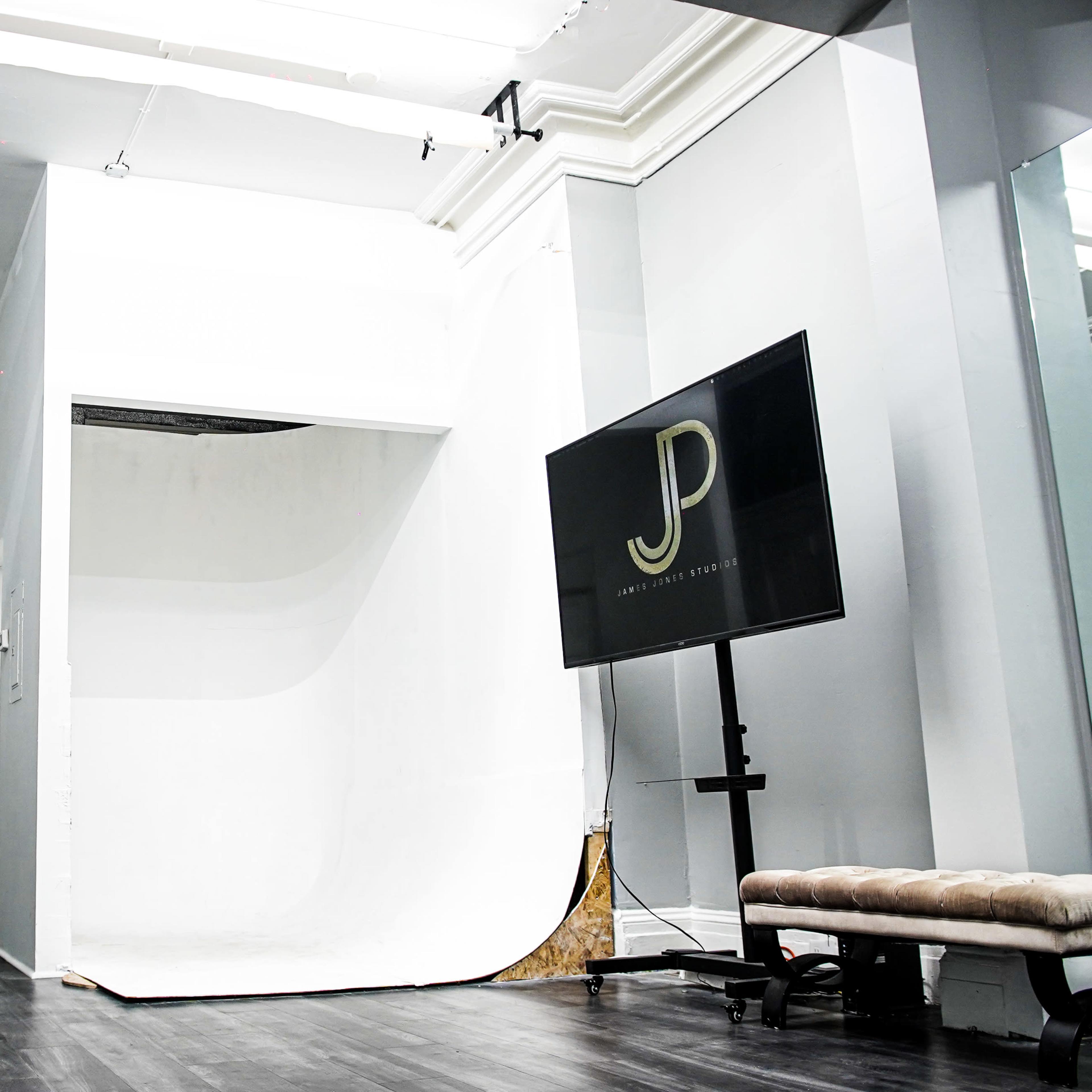A minimalist photography studio with a white backdrop, a large flat-screen television displaying the initials "JP," and studio lighting equipment positioned nearby.