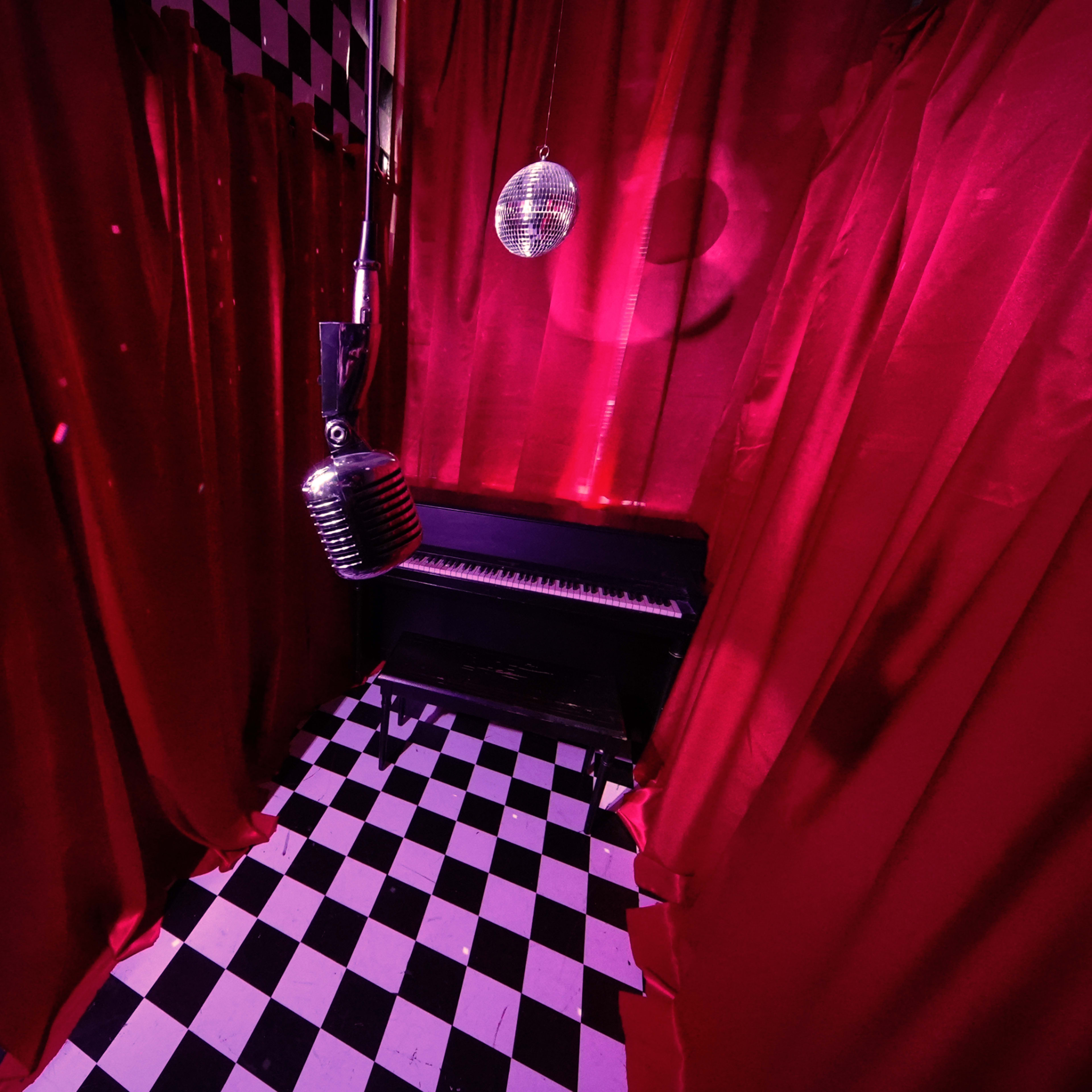 A small room with red curtains, a vintage microphone hanging from the ceiling, a black piano, and a checkered floor.