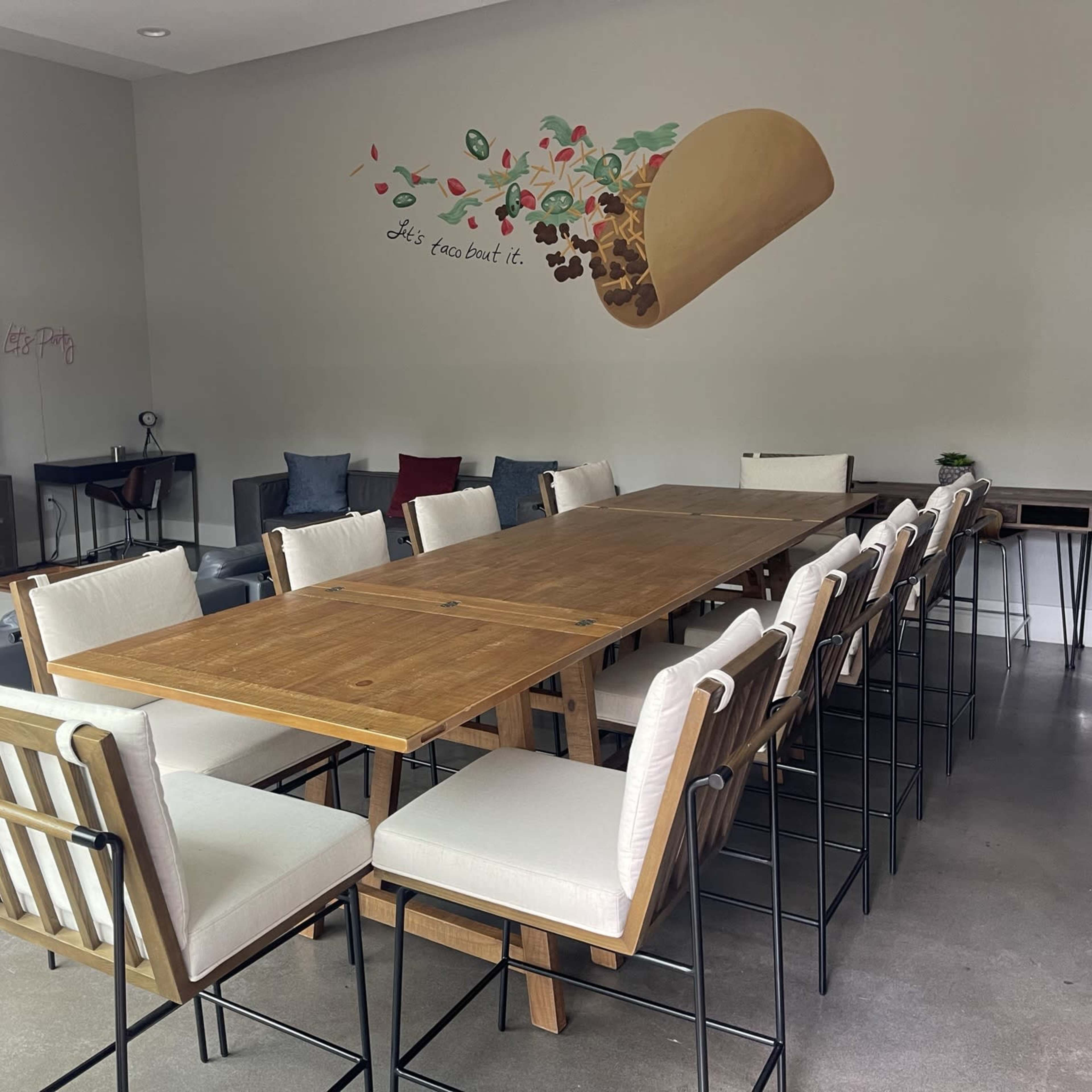 A spacious room with a long wooden dining table surrounded by high-backed chairs, a sectional couch in the corner, a television, and a mural of a taco on the wall.