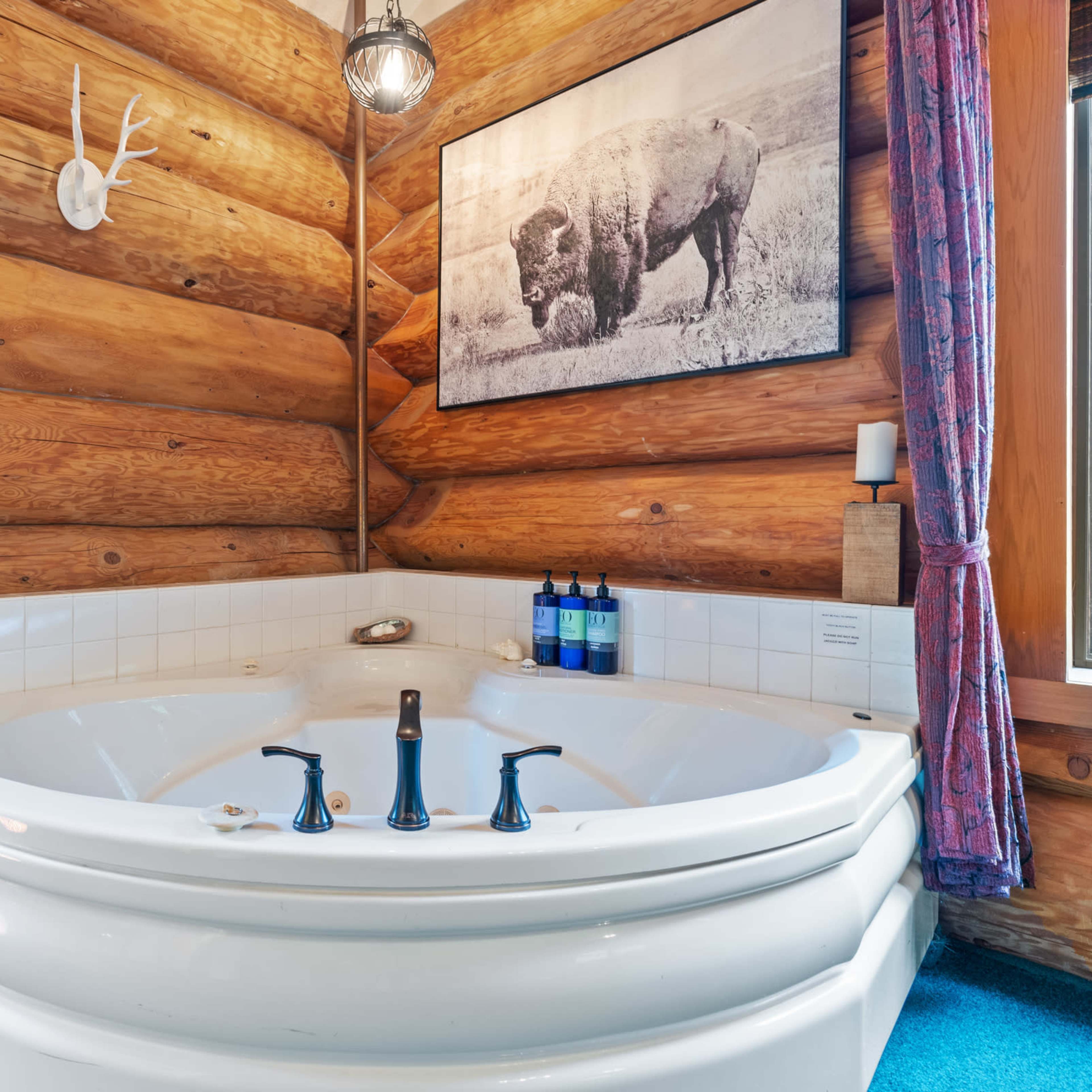 A spacious corner jetted bathtub surrounded by wooden log walls, with a framed buffalo photograph above and candles on the windowsills.
