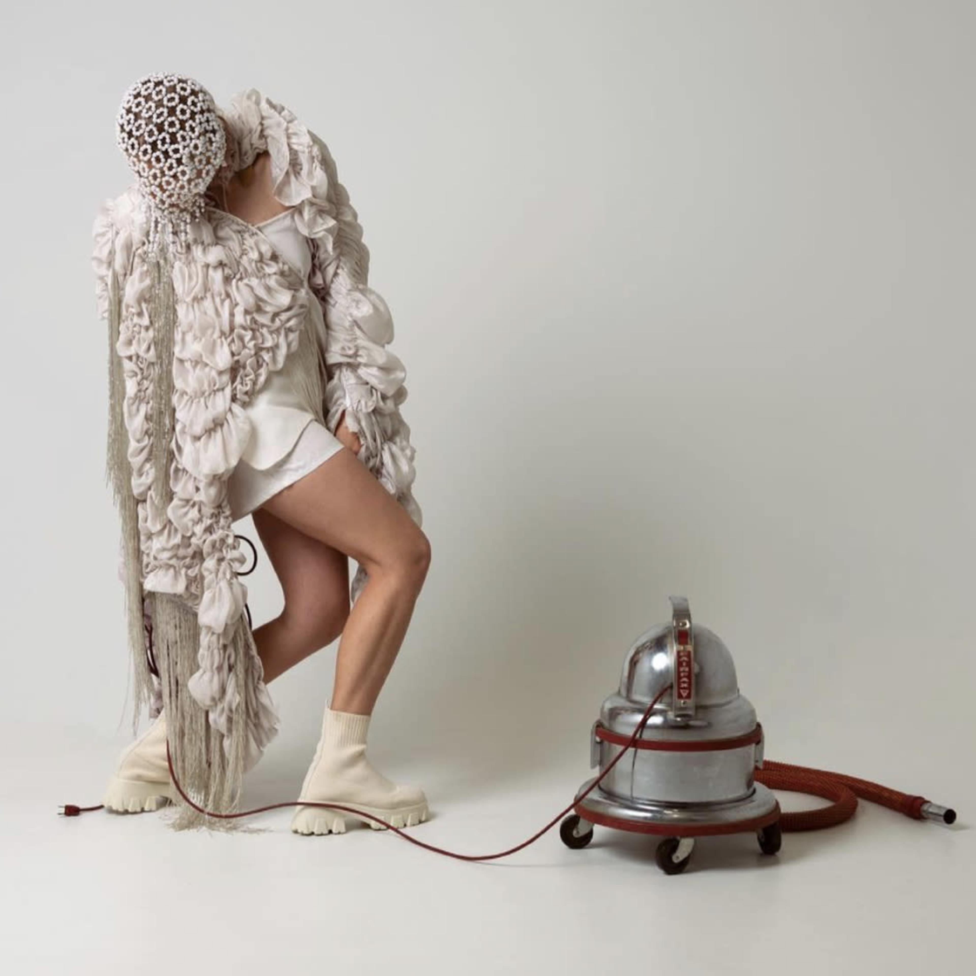 A person in an elaborate textured outfit interacts with a vintage vacuum cleaner in a minimalist setting.
