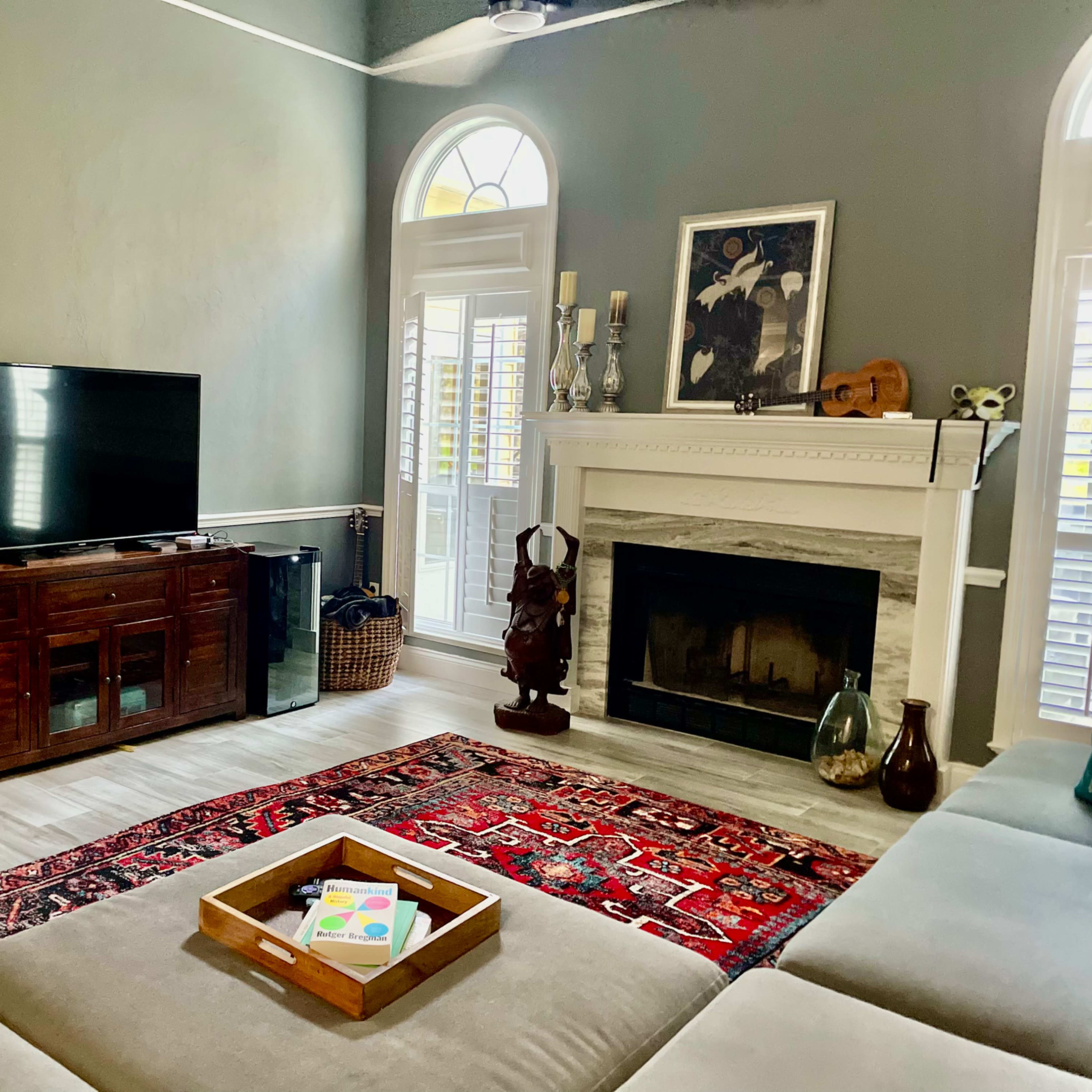 A living room features a television on a wooden cabinet, a stone fireplace, and a rug with a decorative pattern, all surrounded by large windows and natural light.