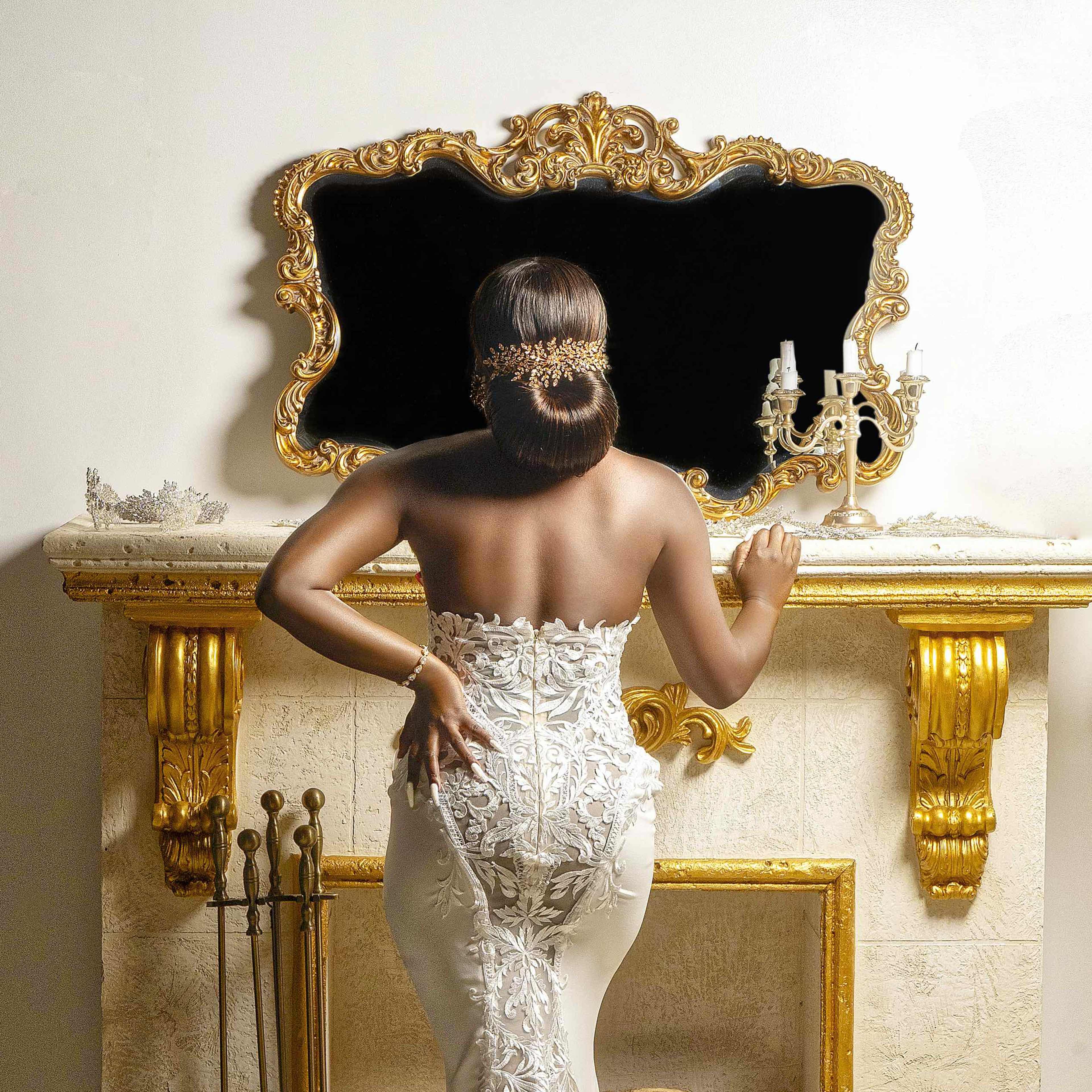 A woman in an ornate white dress stands in front of a gilded mirror above a decorative fireplace.