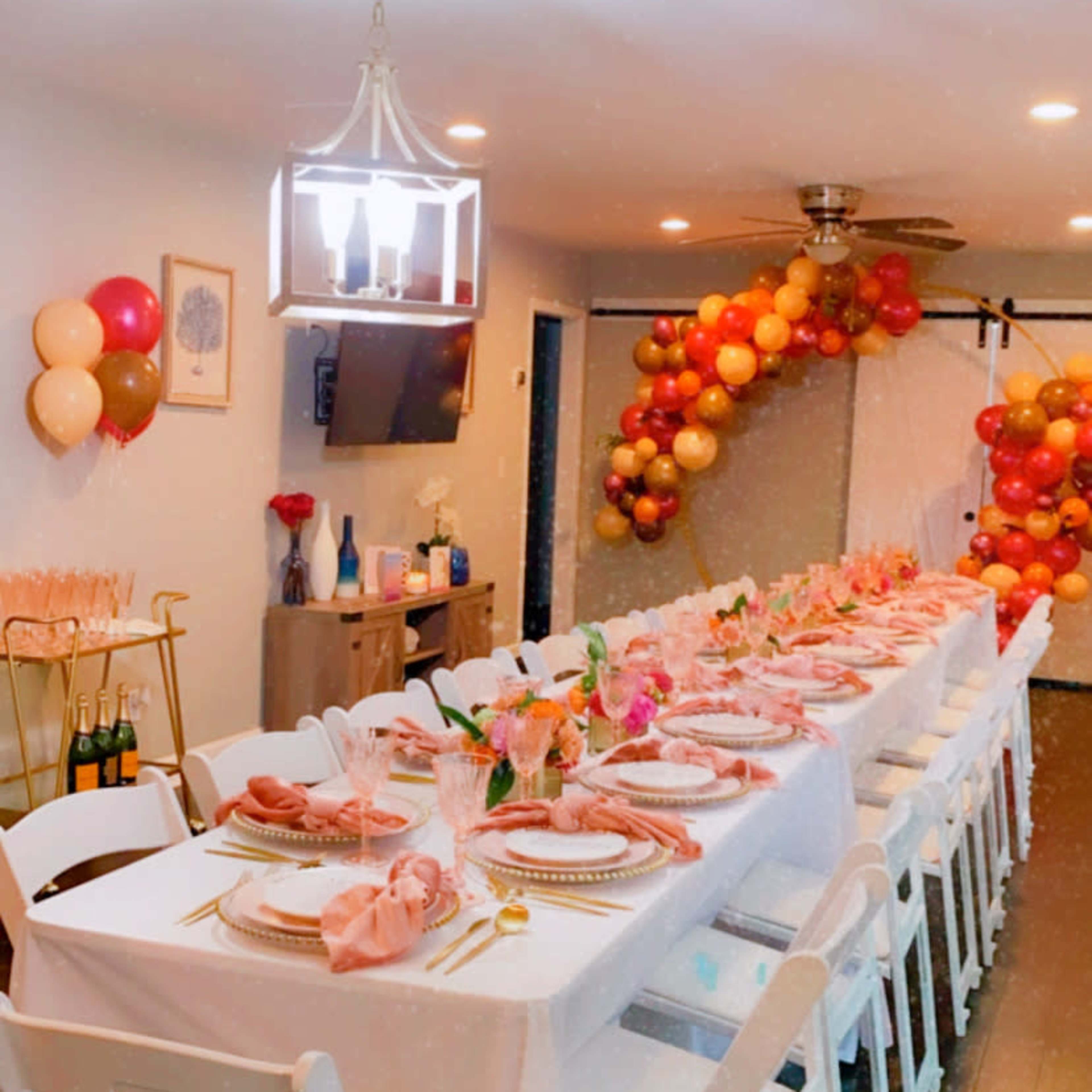 A long white table is set for a celebration, decorated with pink napkins, floral centerpieces, and a backdrop of colorful balloons.