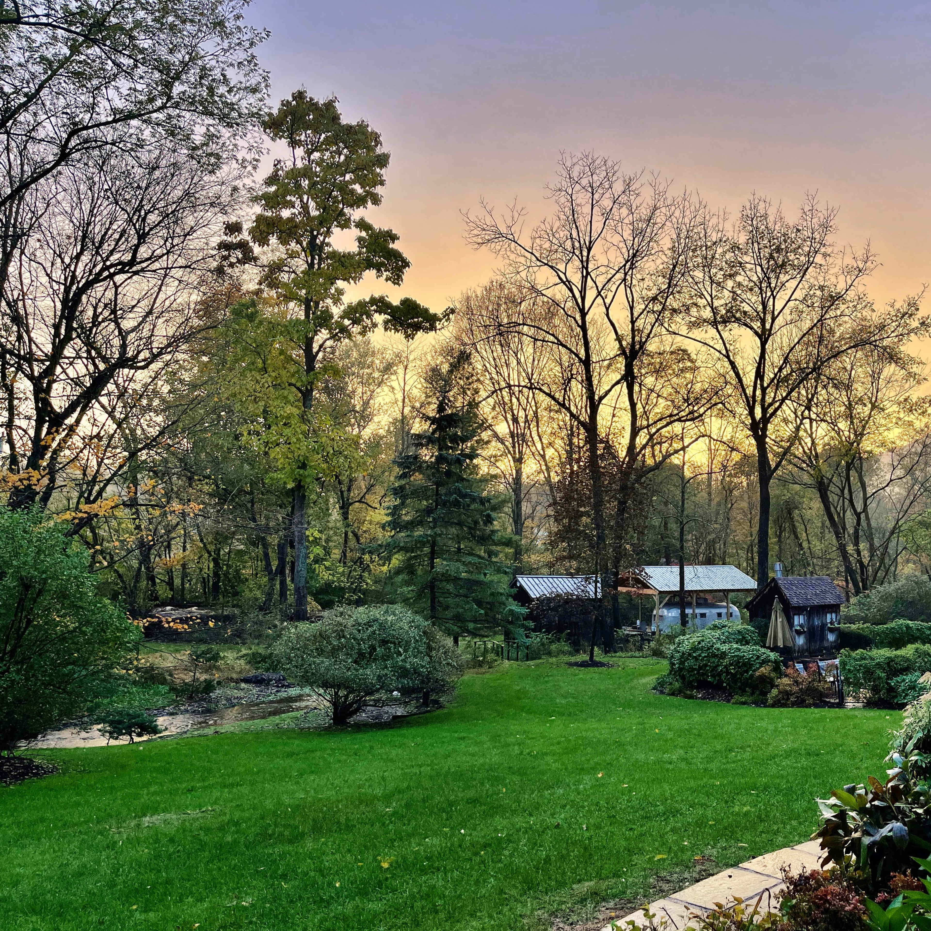 A well-maintained garden with green grass, bushes, and trees, set against a background of wooden structures and a colorful sunset sky.